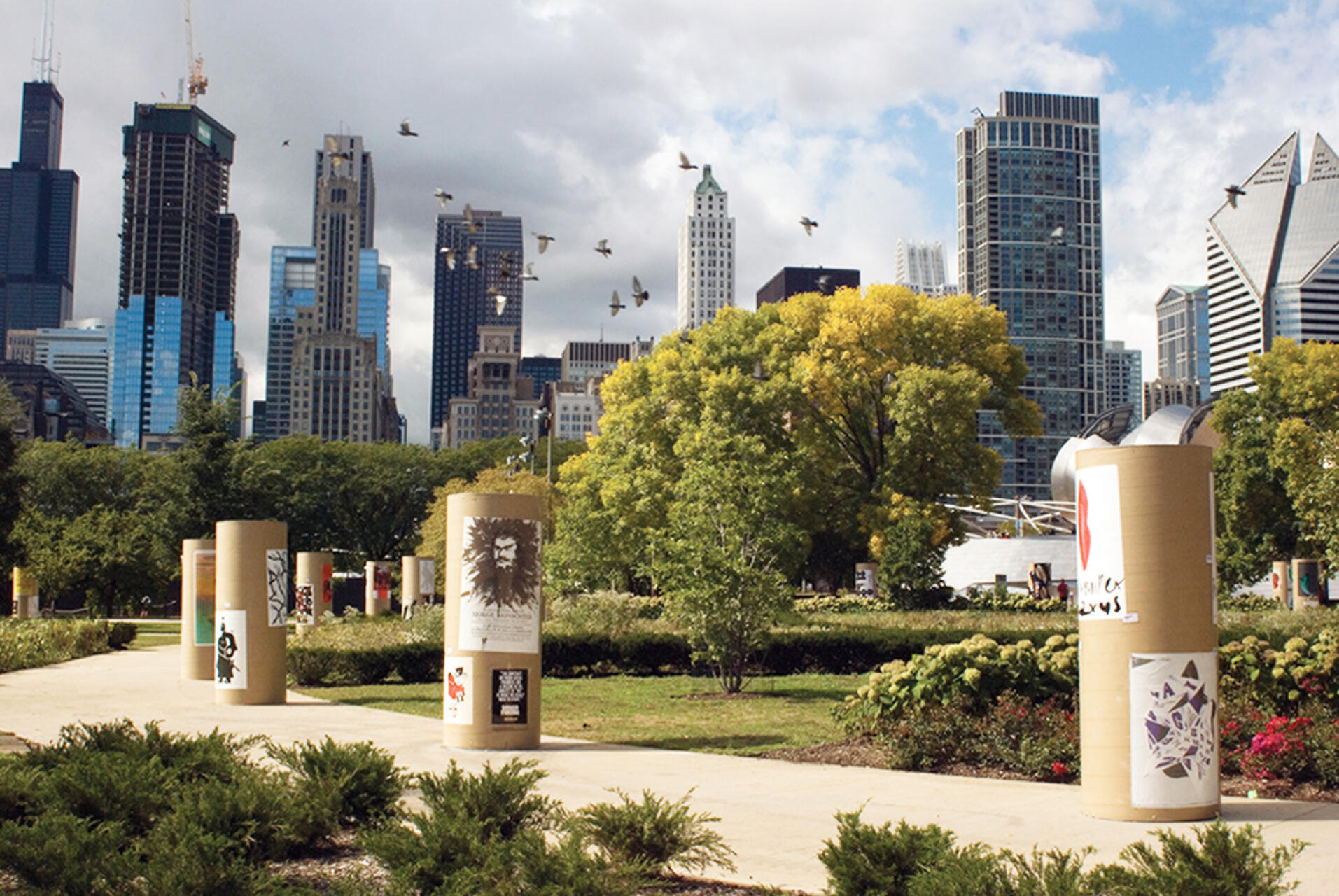 Pathway with decorated columns in a park, surrounded by green trees and shrubs. The city skyline, featuring tall buildings, is visible in the background under a partly cloudy sky, with birds flying above.