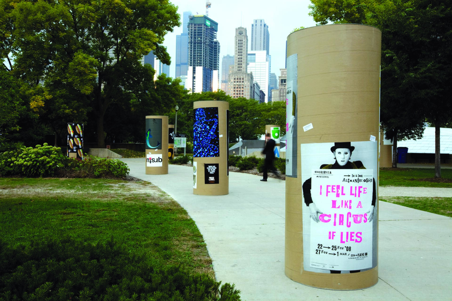 Outdoor park with cylindrical structures displaying colorful posters and graphics. Skyscrapers are visible in the background, surrounded by trees and greenery. A person walks along the path.