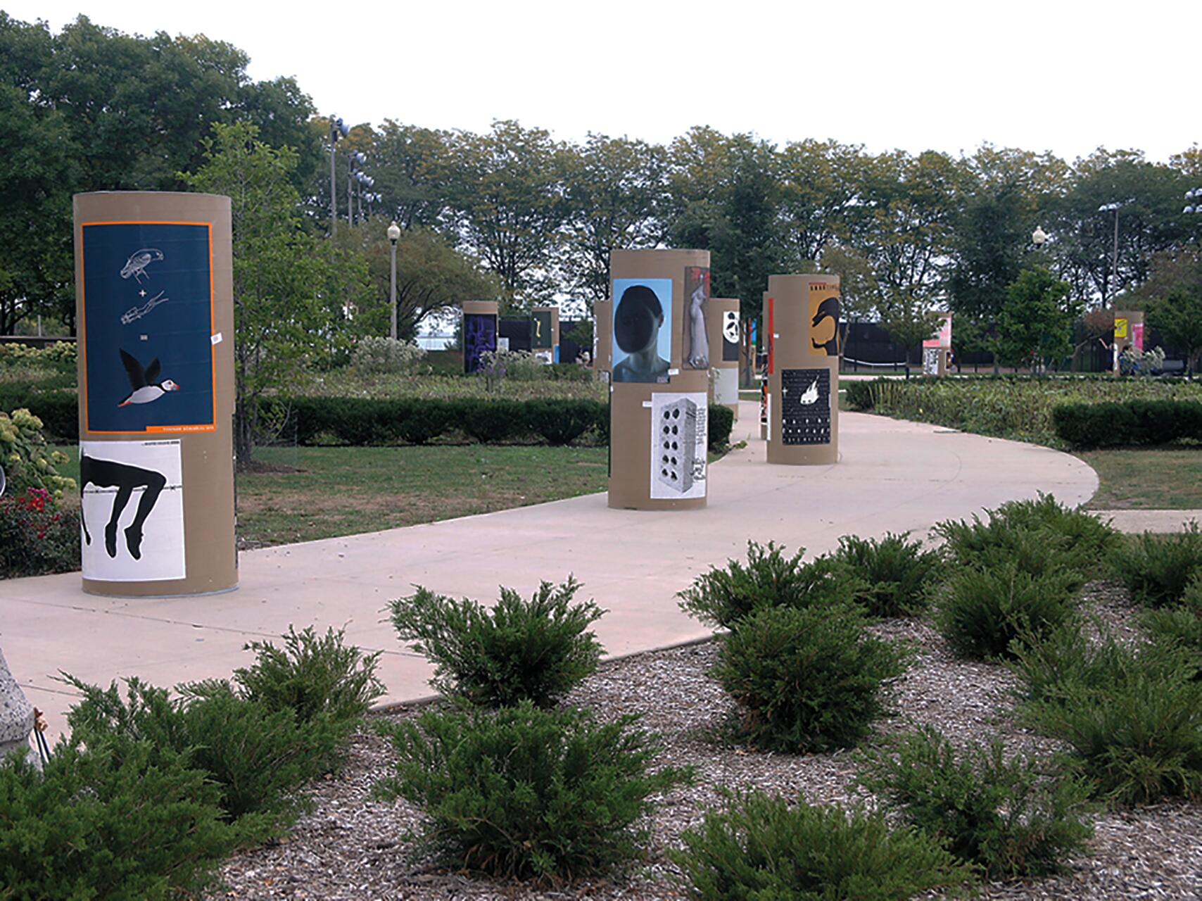 Outdoor park with cylindrical structures displaying various abstract art posters along a curved path, surrounded by greenery and trees in the background.
