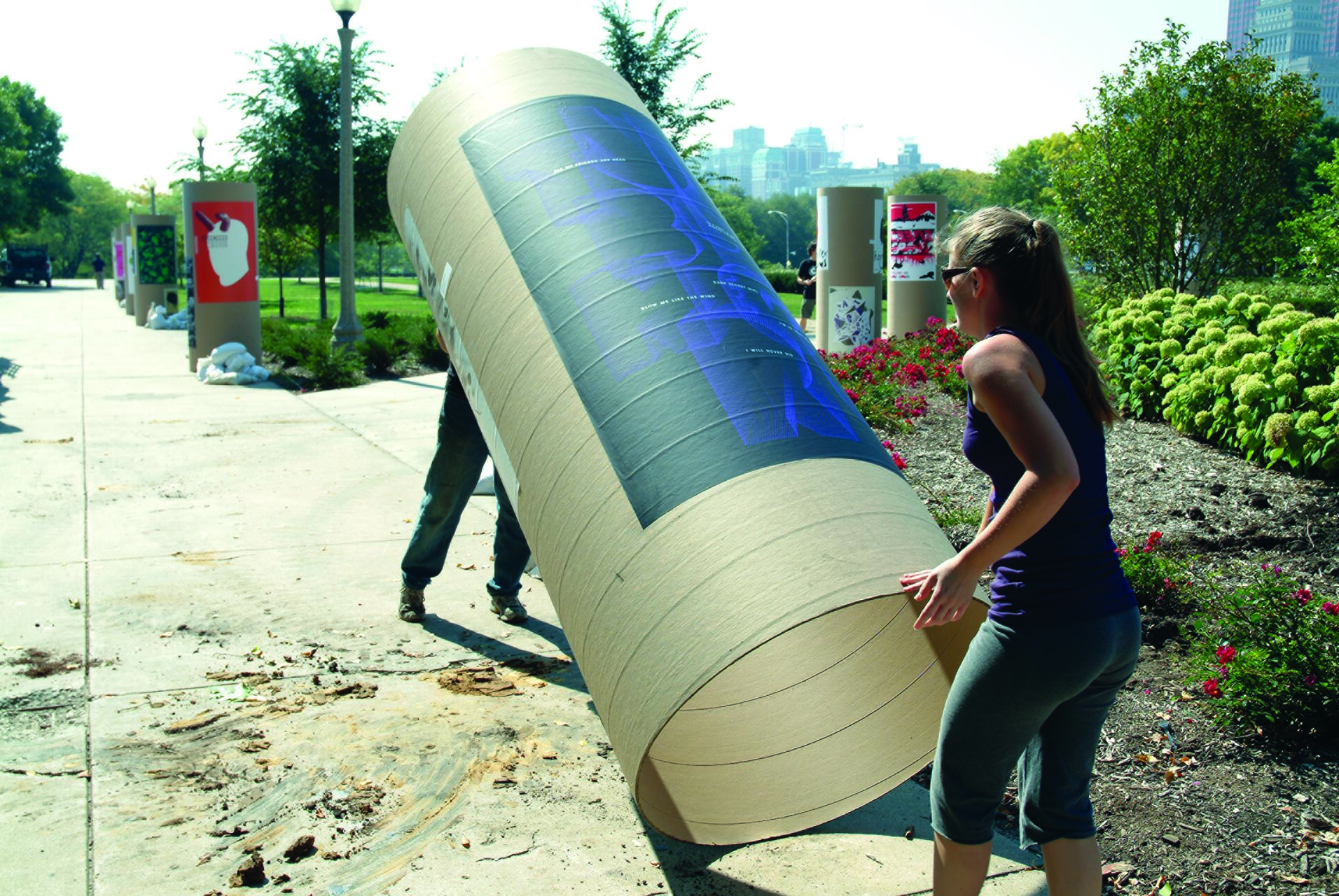 Two people carrying a large cylindrical poster in a park, with blooming flowers and city buildings in the background.