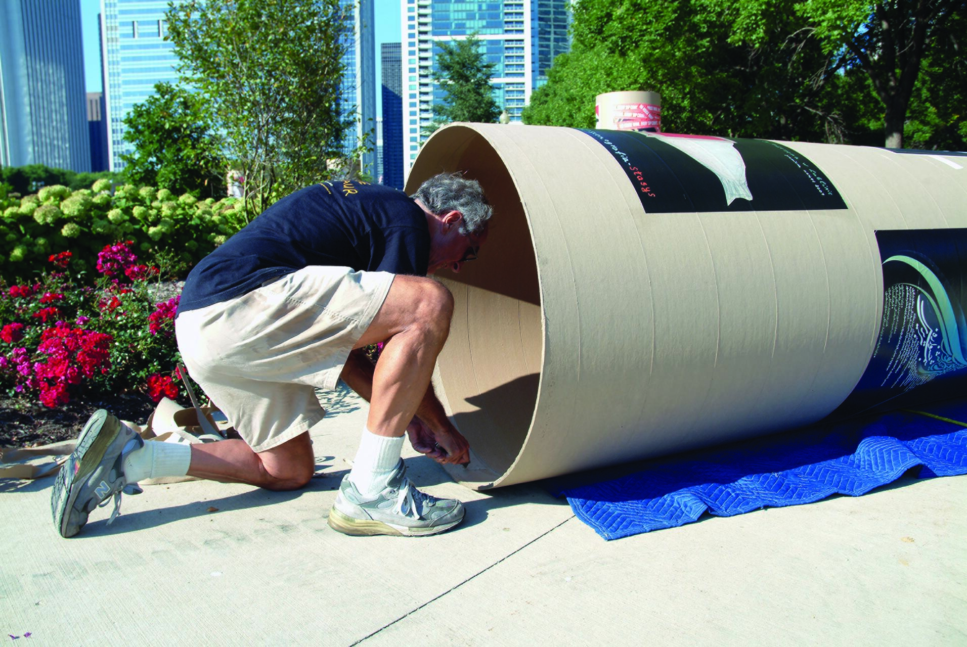 Man wearing shorts and sneakers kneels beside a large cylindrical cardboard structure on a sidewalk in an urban park. He is working on the structure, surrounded by blooming flowers and city skyscrapers in the background.
