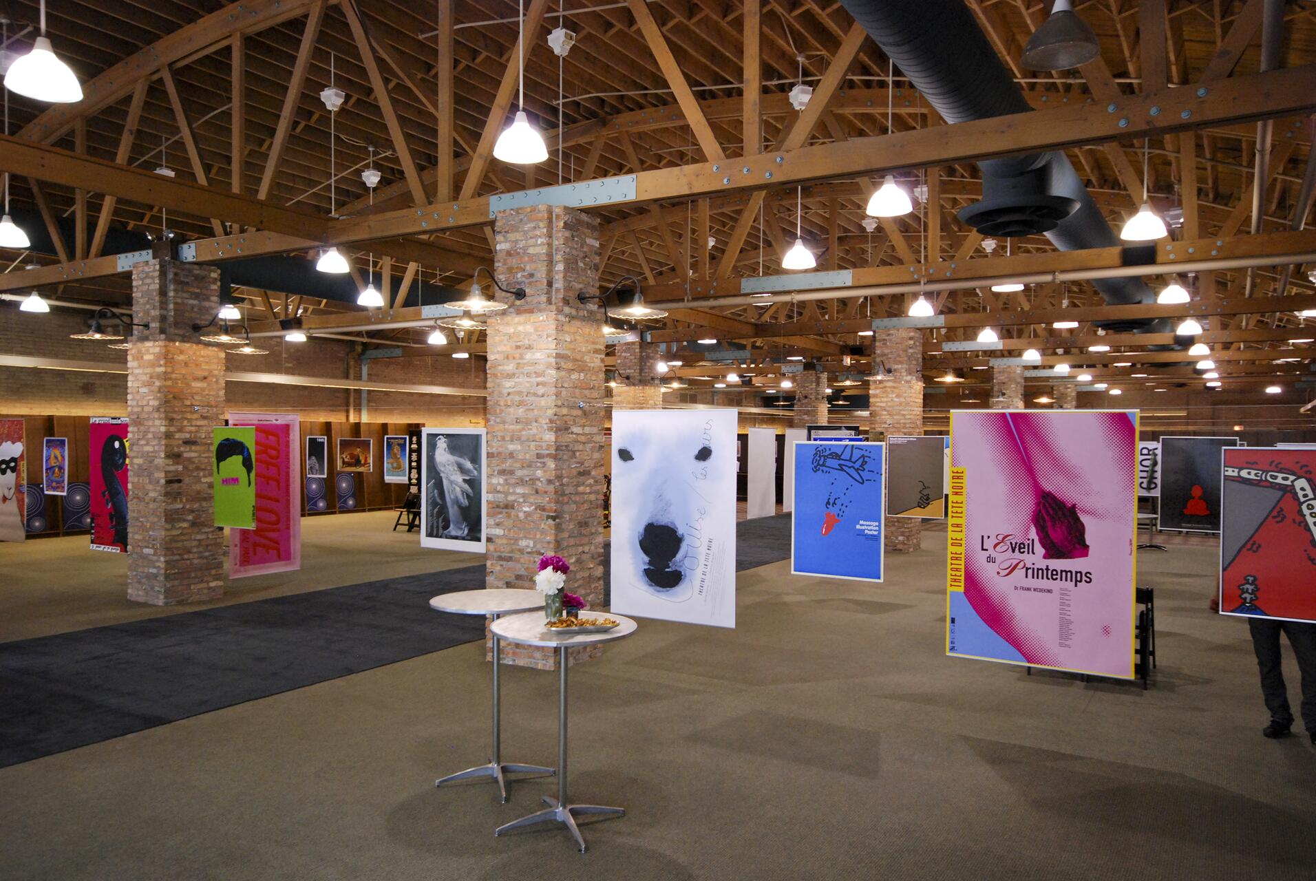 Art exhibition in a spacious gallery with posters and artwork hanging from the ceiling; exposed brick pillars and wooden beams under bright lighting; a small table with flowers in the foreground.