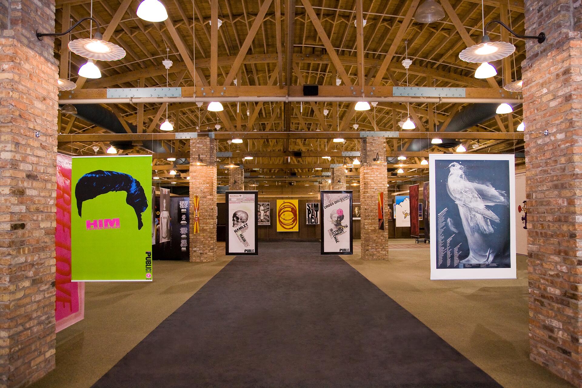 Art gallery interior with exposed brick walls and ceiling beams, featuring colorful modern art posters. A green poster on the left and a blue poster with a bird on the right stand out. Warm lighting creates a cozy atmosphere.