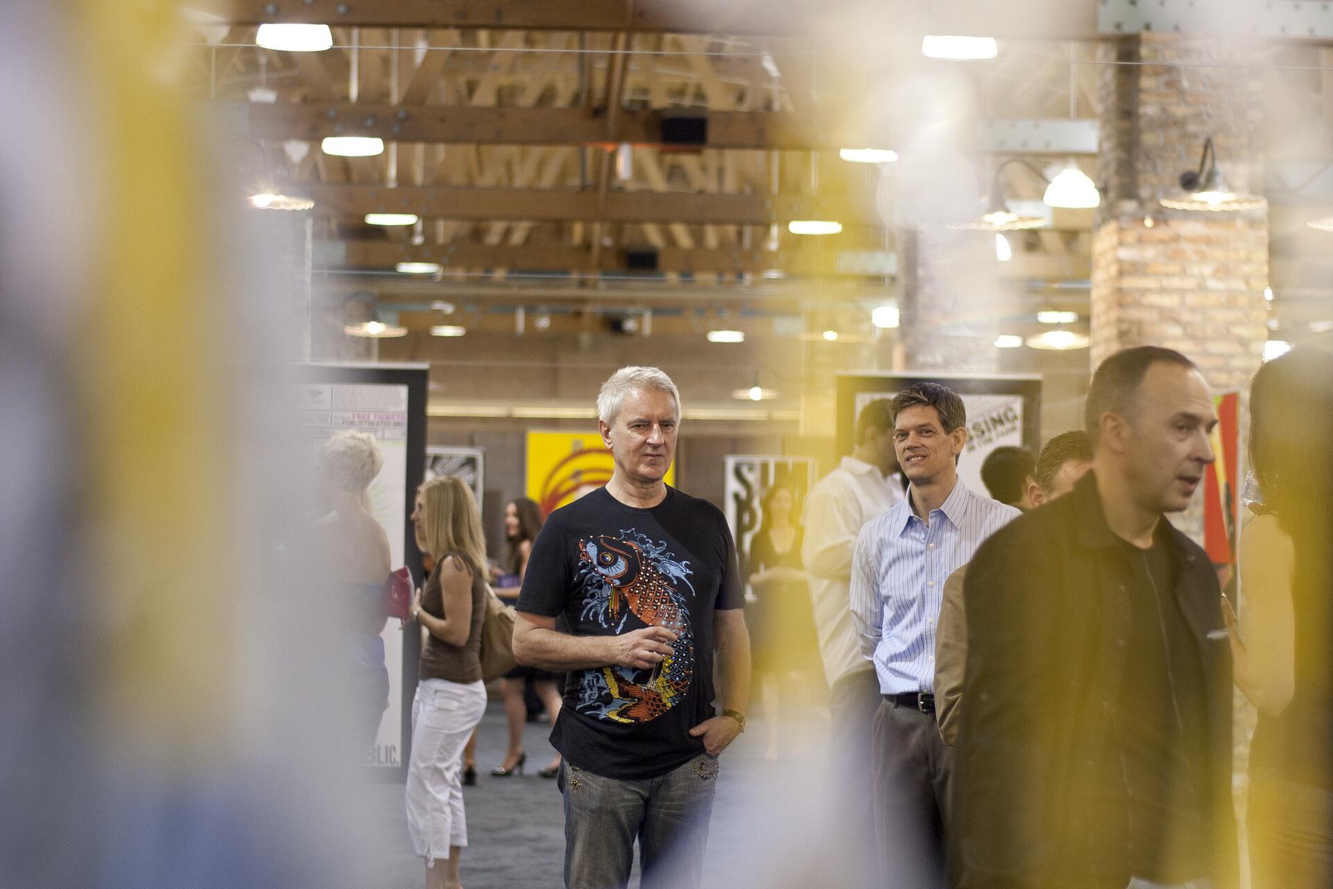 Men and women at an art gallery, with posters on the walls and a wooden ceiling. A man in a dark graphic t-shirt is in the foreground, and other attendees are visible in the background.