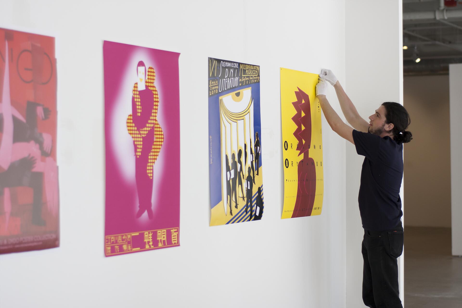 Man with black hair and gloves hangs colorful posters on a gallery wall, including a pink and yellow graphic design.