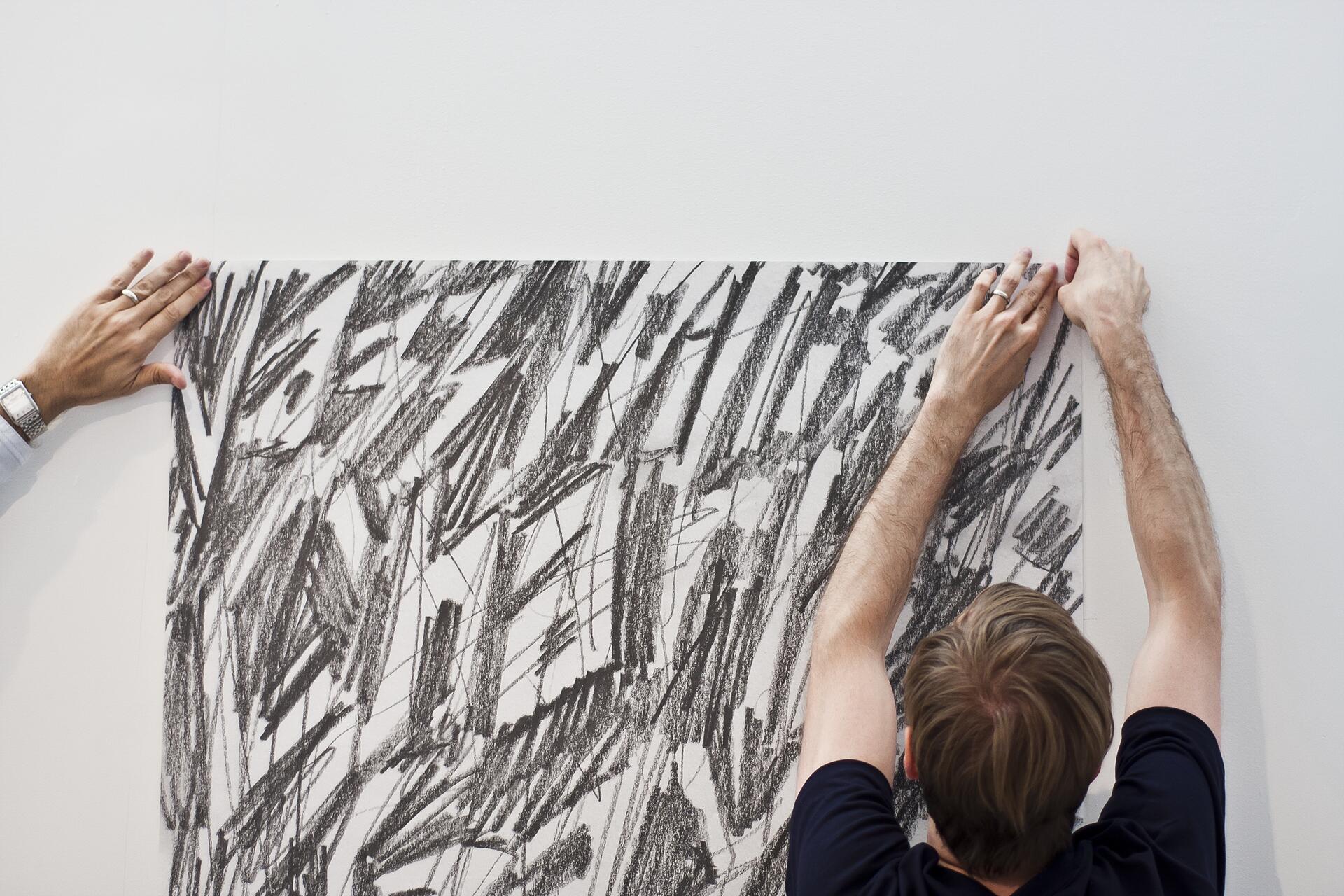 Two people hang a large, abstract black and white drawing on a white wall, each holding a corner.