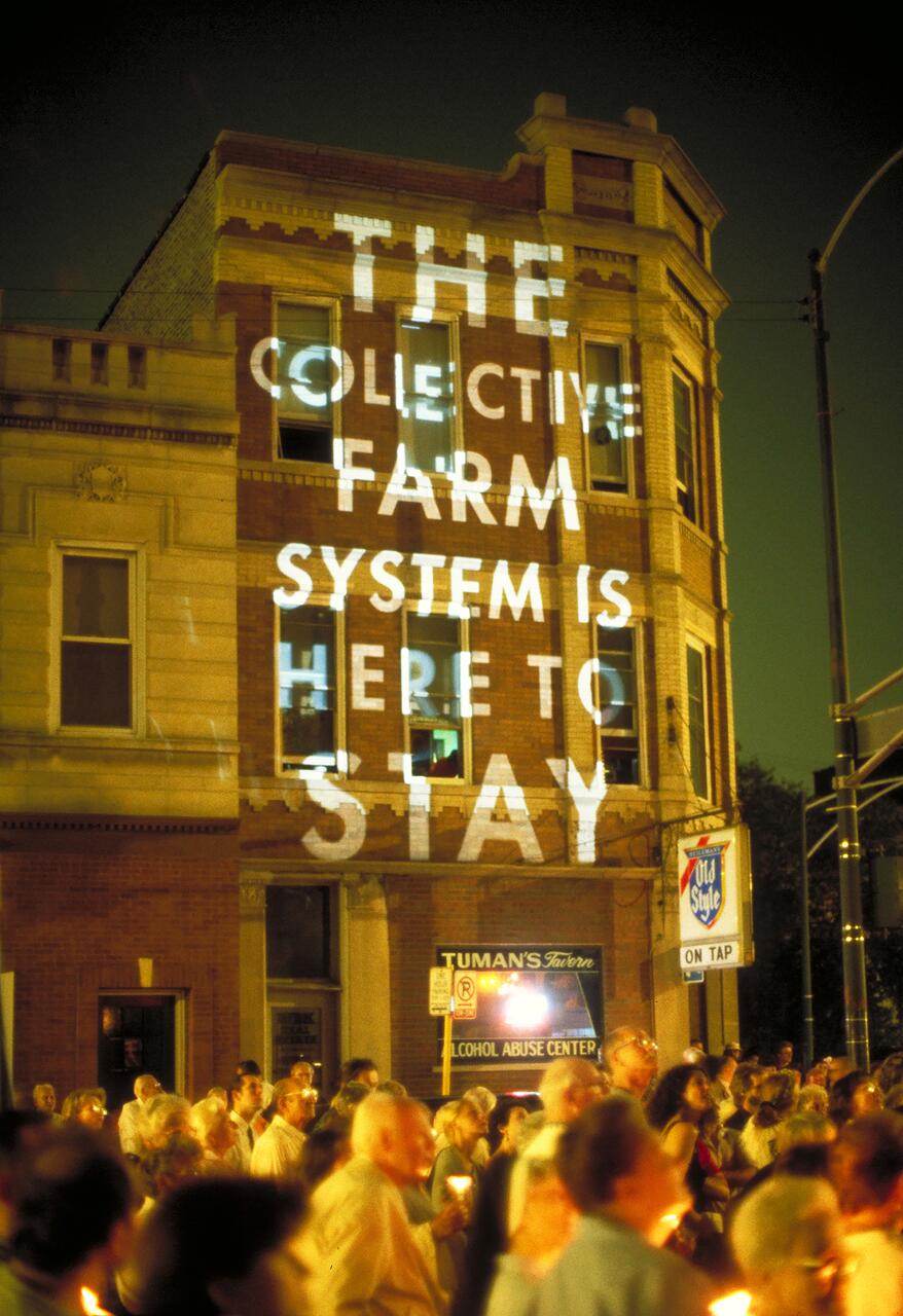 A group of people holding candles gather at night in front of a brick building. Large white text is projected onto the building's facade.