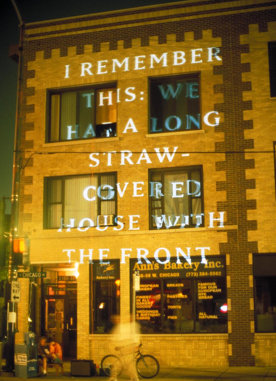 Text projected onto a brick building facade at night, displaying a message about a straw-covered house. Below, Ann's Bakery is visible with illuminated signage and a storefront window, with a blurry person and bicycle nearby.