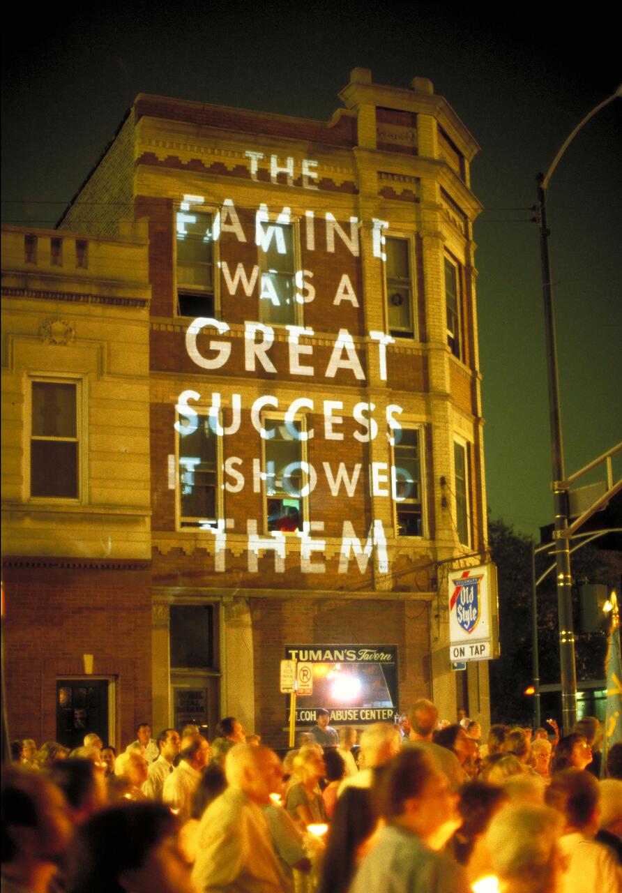 A nighttime street scene with a crowd holding candles outside a brick building. Large, bold white text is projected onto the building's facade.