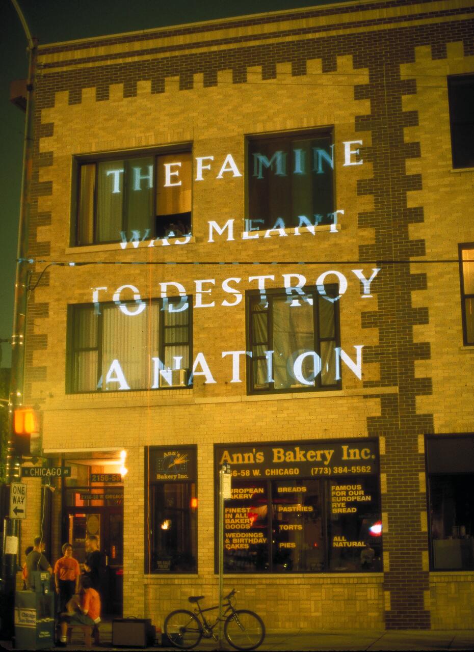 Text projected on a brick building reads "The Famine was meant to destroy a nation." Below, Ann's Bakery Inc. is visible, with a bicycle parked outside and people standing near the entrance.