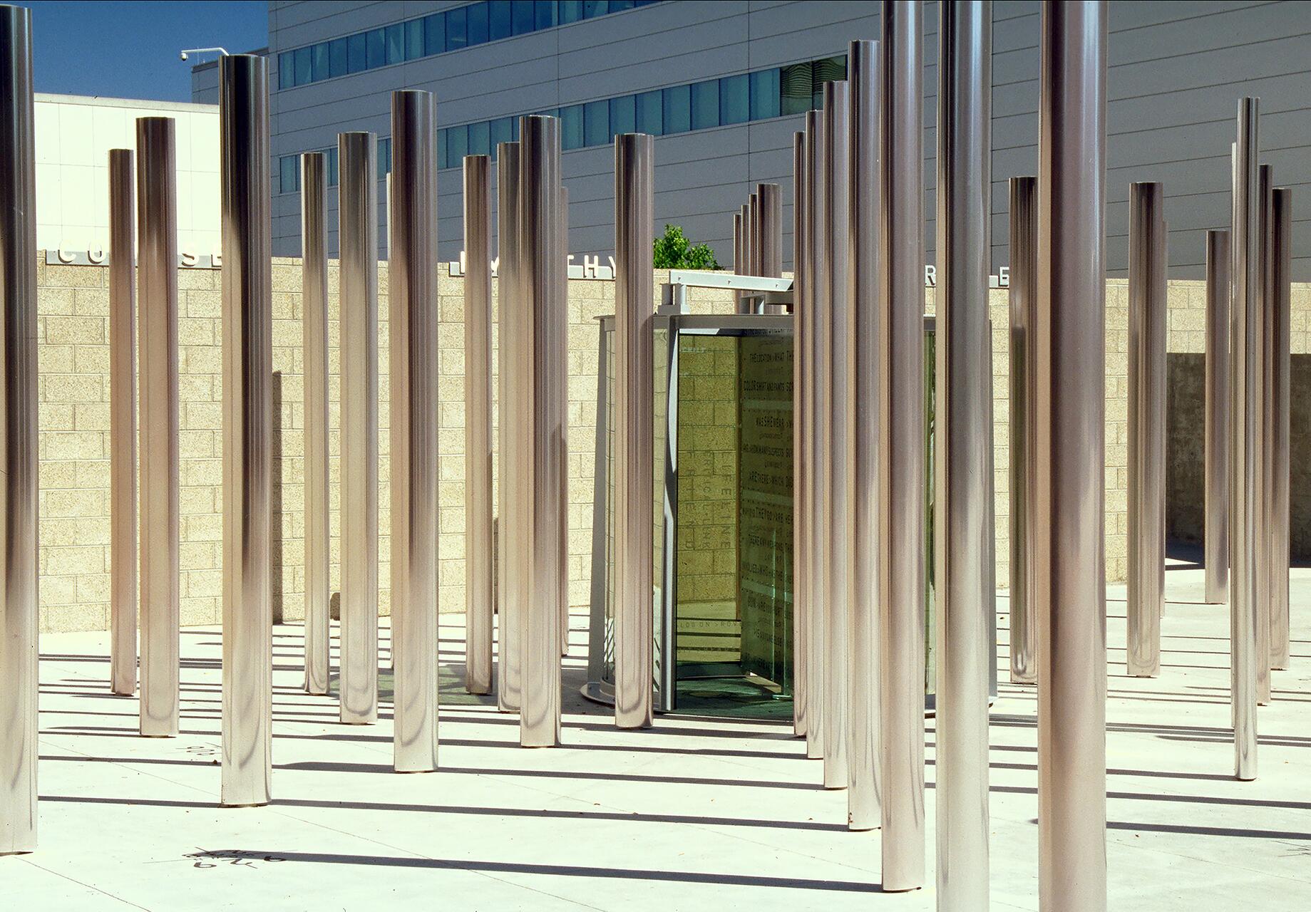 Tall, reflective metal columns are arranged in a grid pattern on a concrete surface. Behind them, a modern wall with windows and a glass panel door is visible, casting distinct shadows on the ground.