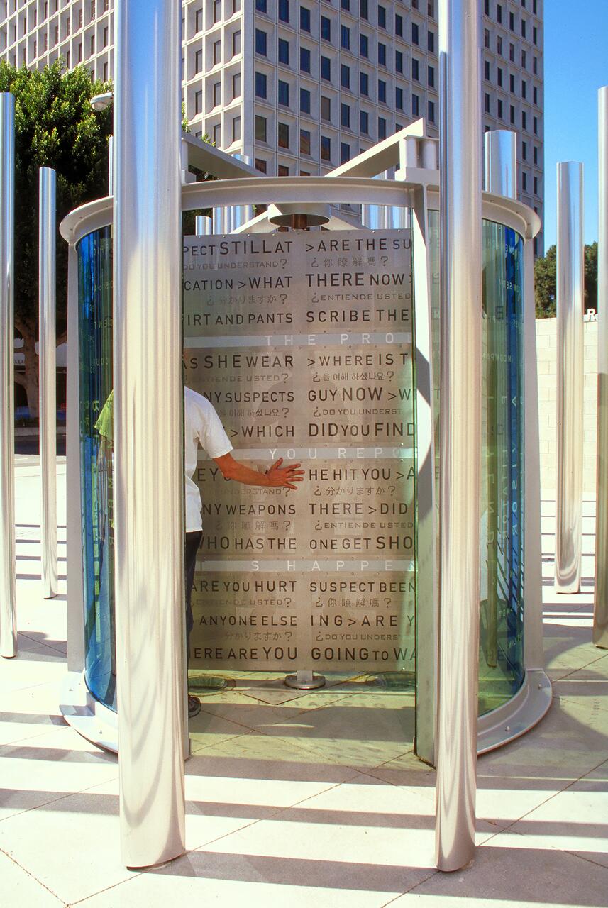 Man standing inside a modern, cylindrical sculpture made of glass and metal, partially obscured by the structure. The sculpture has etched text in multiple languages on its surface. A tall building and trees are visible in the background.