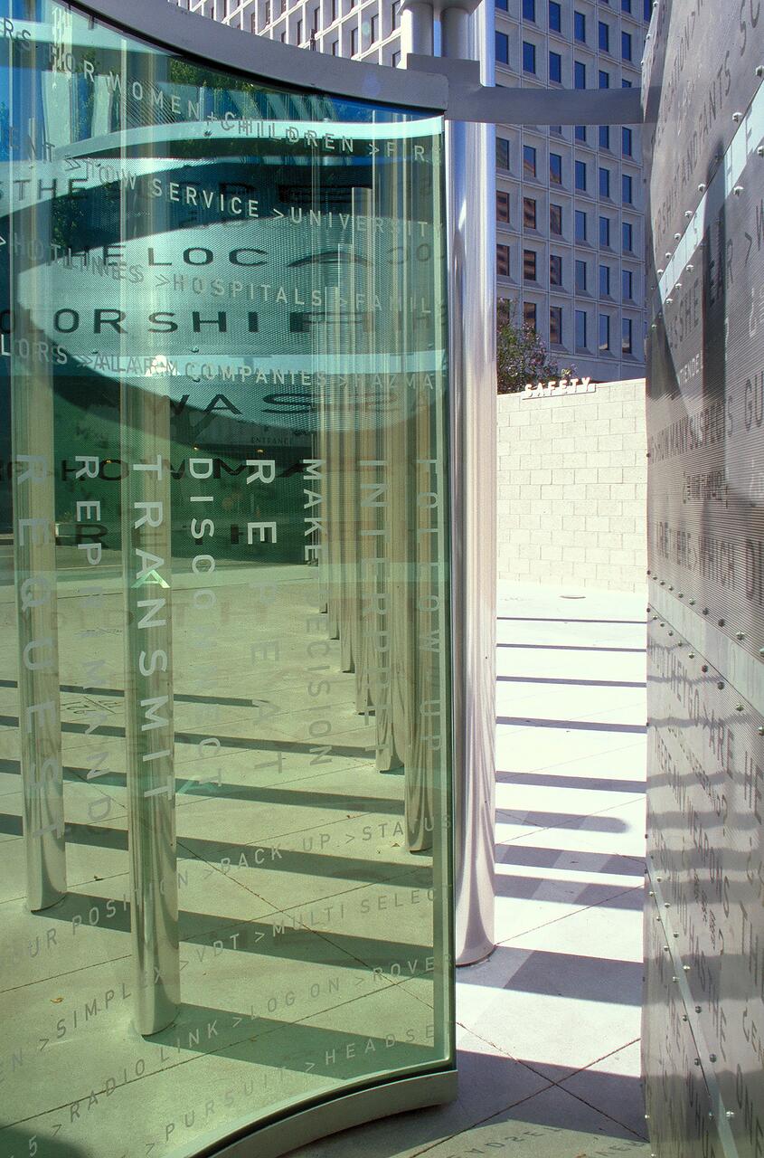 Curved glass panels with etched text, reflecting a modern building in the background. Metal columns and shadows on the pavement create a geometric pattern.