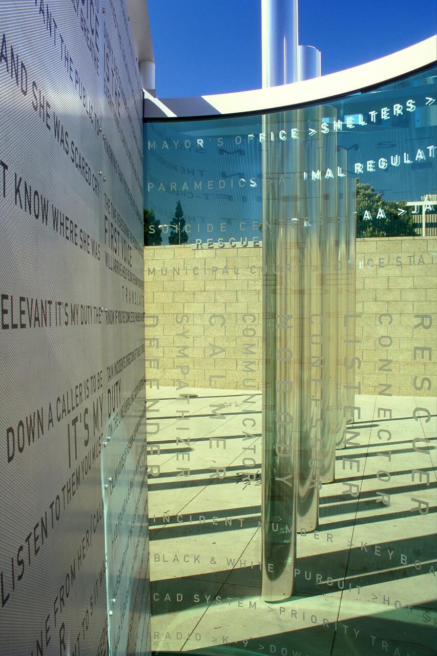 Reflective metal columns and glass panels with etched text, casting sharp shadows on the pavement, set against a clear blue sky and brick wall.