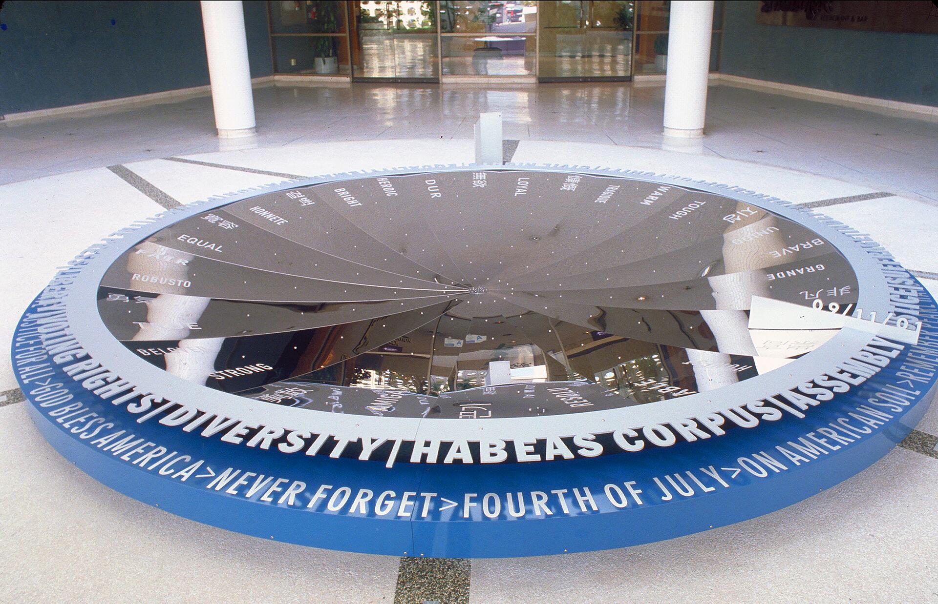 Large reflective sundial with various words engraved on the surface, encircled by a blue band with phrases like "God Bless America" and "Fourth of July." The setting is indoors with a polished floor and large windows in the background.