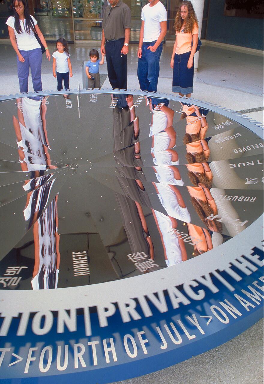 People standing around a large reflective circular sculpture, with text and distorted reflections visible on the surface, in a modern indoor setting.