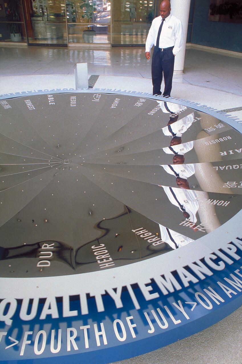 A man in a white shirt and dark pants stands next to a large reflective sculpture on the floor, showcasing words like "EQUALITY," "EMANCIPATION," and "FOURTH OF JULY." His reflection and additional words are visible on the shiny surface.