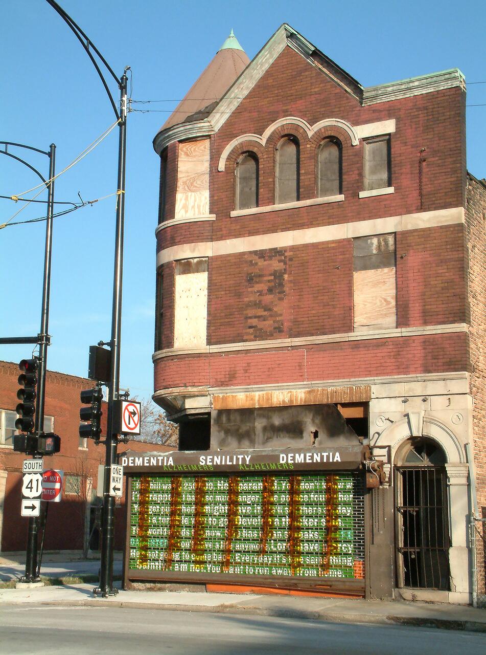 Old brick building with boarded windows, graffiti on the lower facade, and a vintage style turret. Traffic lights and street signs are visible in the foreground, while the sky is clear and blue.