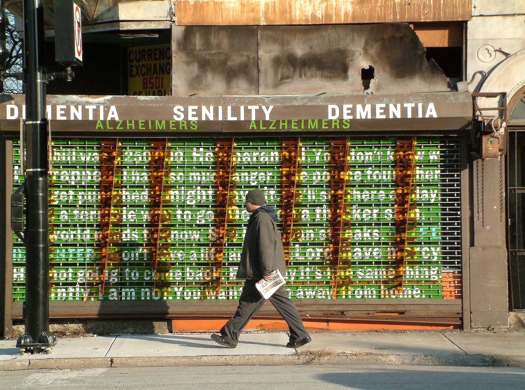 Man in a coat and hat walks past an art installation with words like “dementia” and “Alzheimers” above a grid with layered text in green and orange, on a city sidewalk. He carries a newspaper.