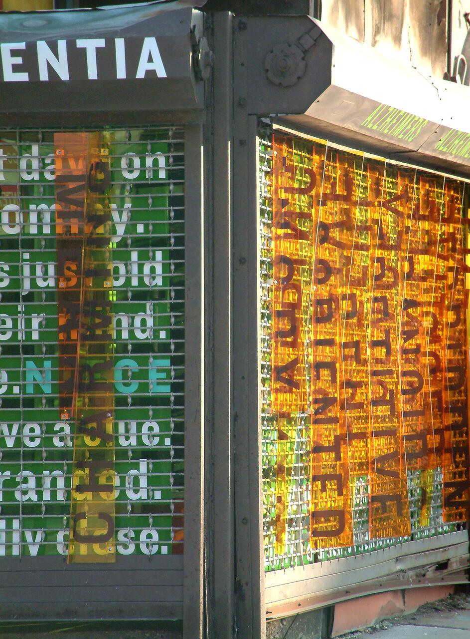 Colorful panels with layered text in various fonts and sizes, overlapping each other, set against a wire grill. Orange and green translucent strips create a vibrant effect. The setting appears to be an urban street environment.