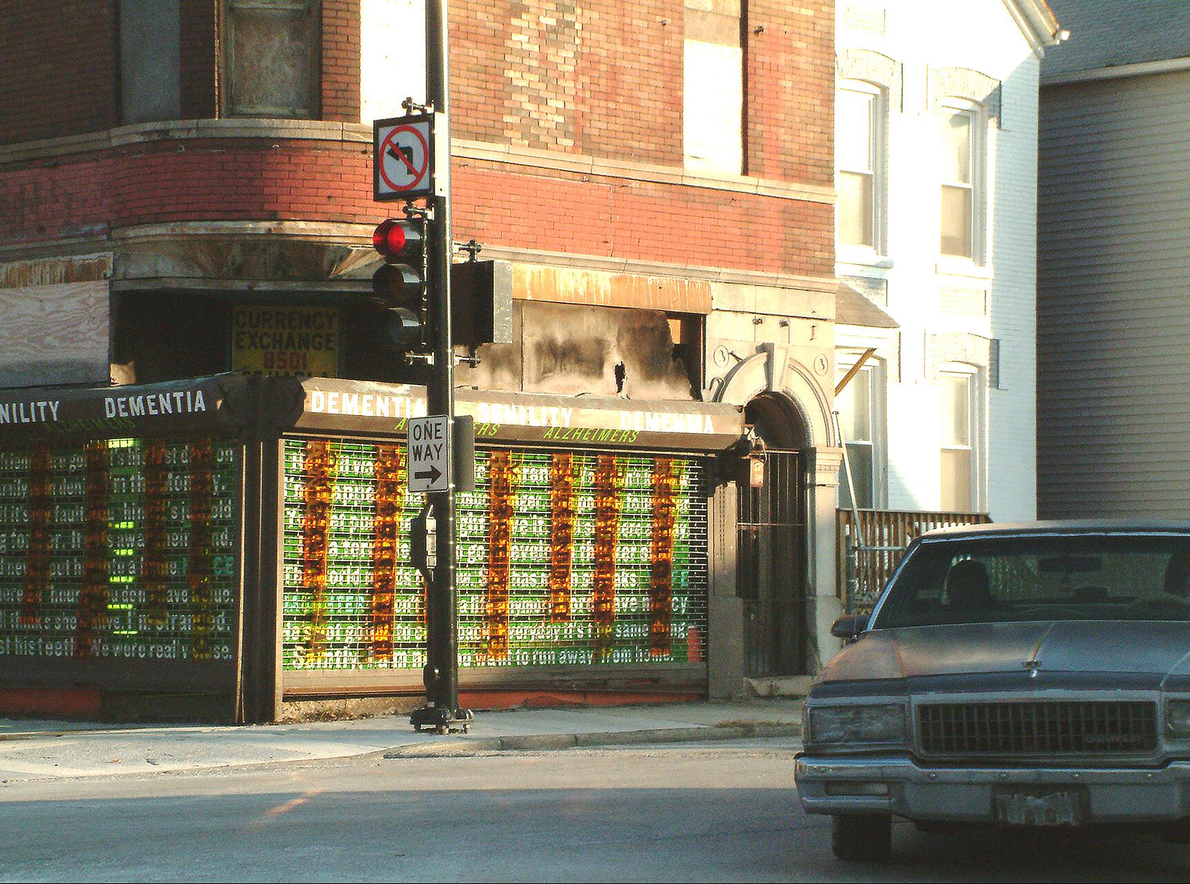 Corner building with red brick, large windows covered with letters and words in various colors. A one-way sign and a red traffic light are on the corner. A vintage car is parked on the street.