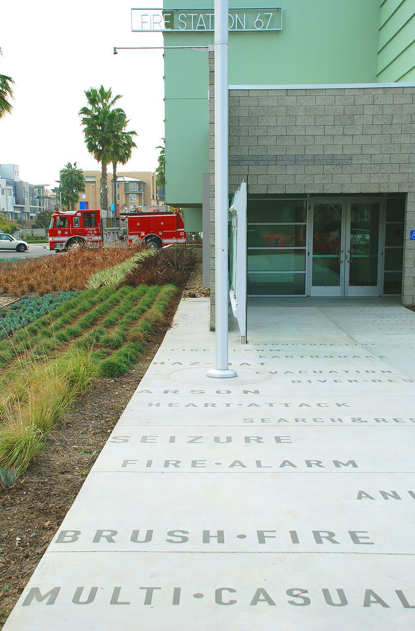 Fire Station 67 entrance with concrete pathway featuring engraved emergency-related words. A red fire truck is parked beside the station. The setting includes palm trees and modern buildings in the background.