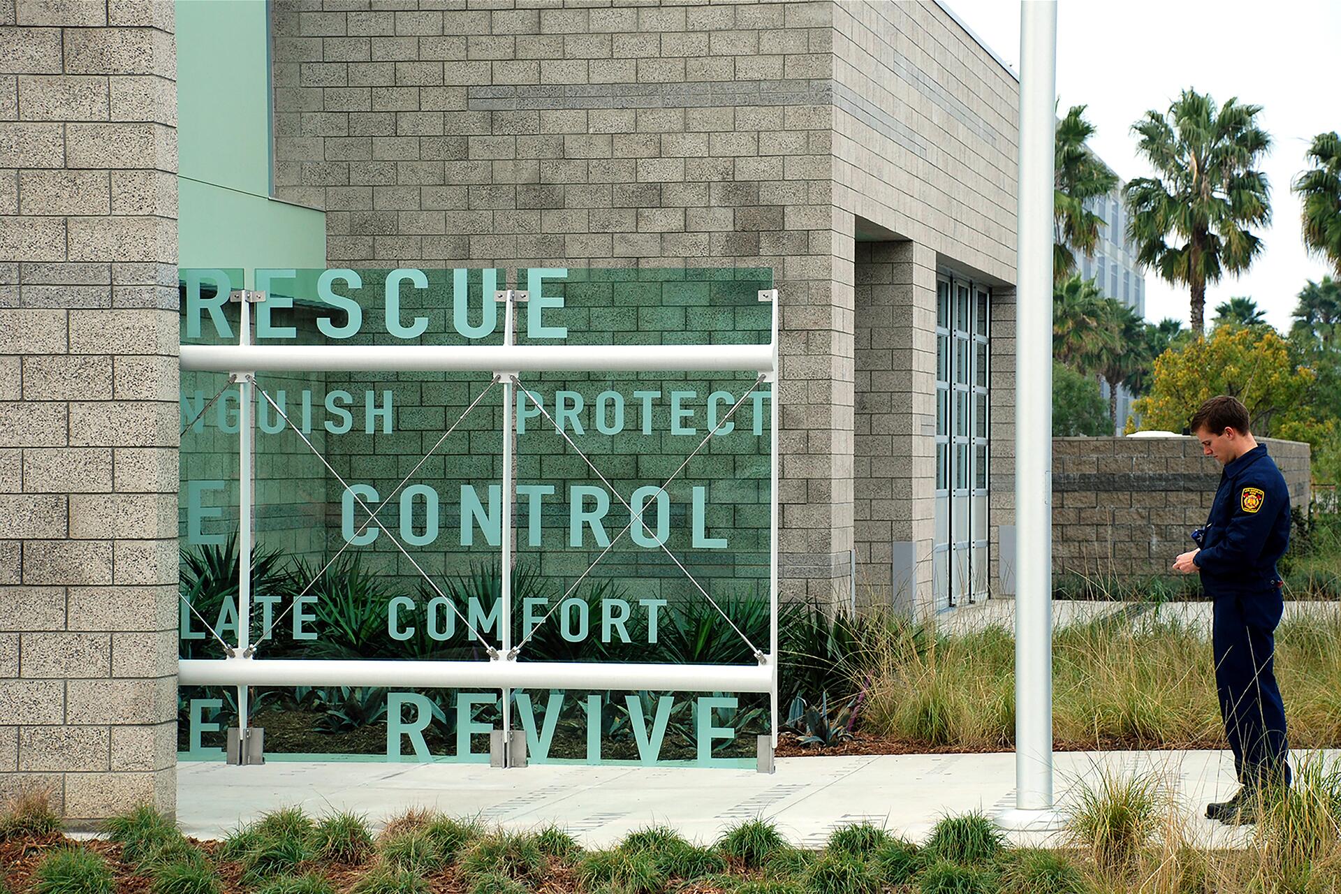 A firefighter in uniform stands outside a building with large words like "RESCUE" and "CONTROL" displayed on a green and white sign. The building is surrounded by palm trees and greenery.