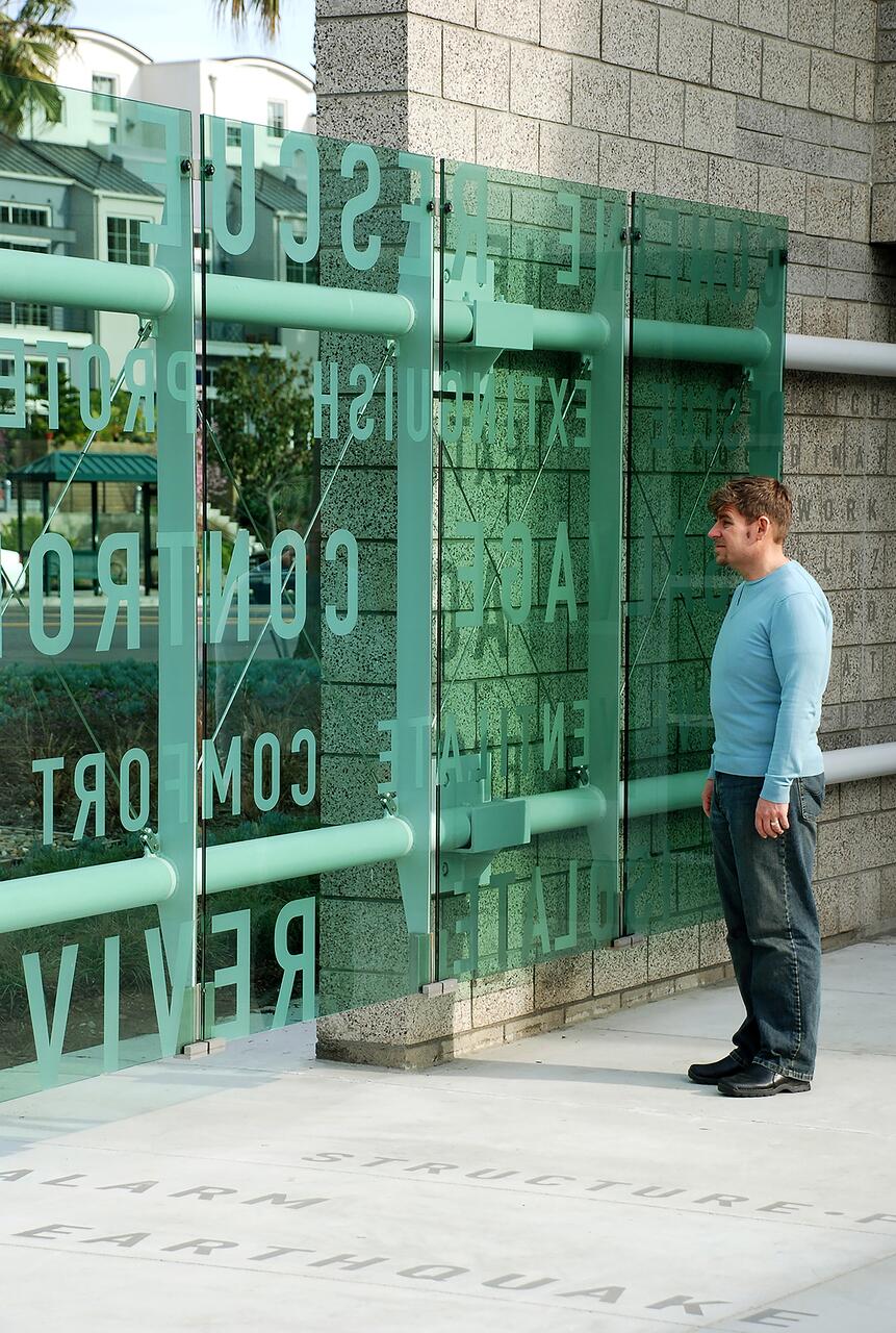 Man in a blue shirt stands facing a glass wall with text, next to a stone building corner.