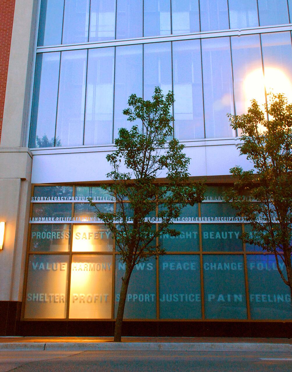 Glass building facade with large windows reflecting a glowing light. Words like "Progress," "Safety," and "Harmony" are visible on lower windows. Two trees stand in front of the building, with a soft evening light illuminating the scene.