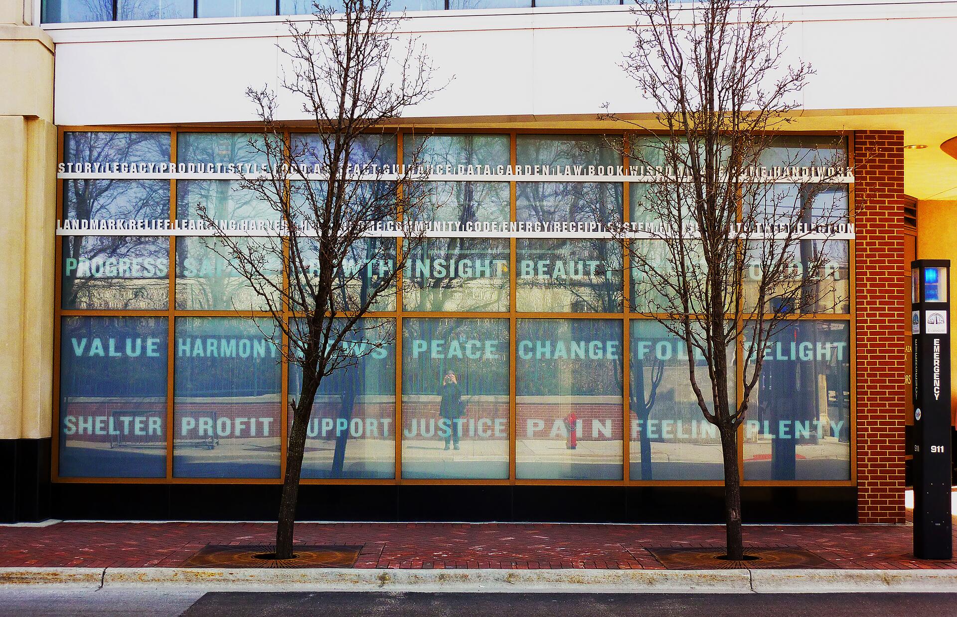 Large windows reflecting trees and displaying words like "progress" and "harmony" in a grid pattern on a brick and concrete building facade. Two leafless trees and a sidewalk in the foreground.