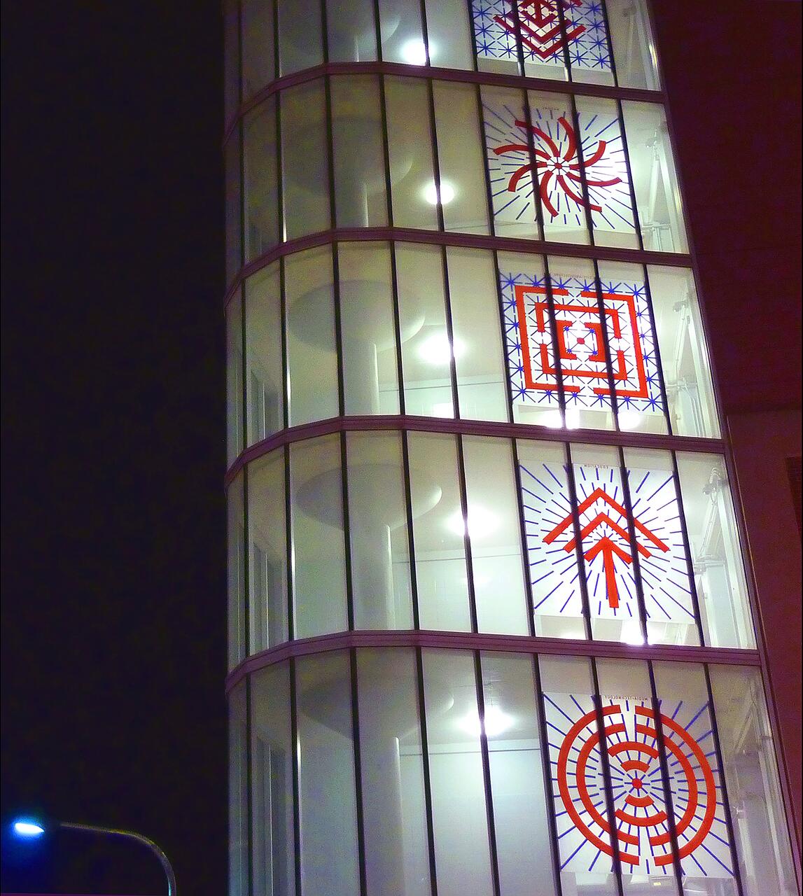 Curved glass building facade at night with geometric red and blue designs on the windows, illuminated from inside.