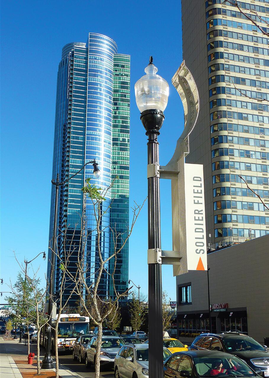 Street with bare trees and cars parked alongside. A tall lamppost features a "Soldier Field" banner. Modern high-rise buildings loom in the background against a clear blue sky.