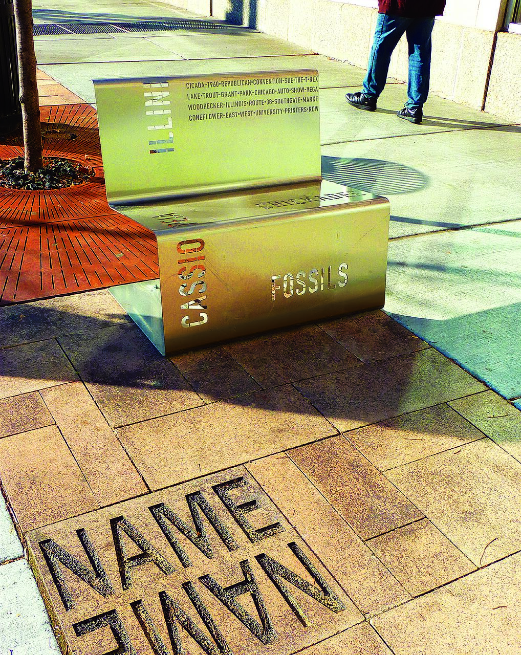 Sculptural street bench with perforated text, located on a sunny sidewalk. A person wearing jeans and black shoes walks nearby. "NAME" is carved into the pavement in front of the bench.