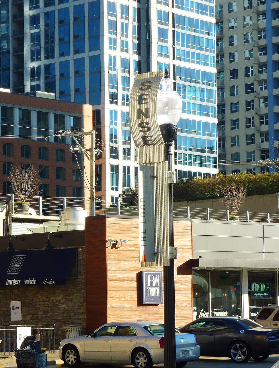 Street scene with tall buildings in the background. A lamppost displays the word "SENSE." Below, a storefront with the sign "URBAN COUNTER" offers burgers, sandwiches, and shakes. A couple of cars are parked nearby.