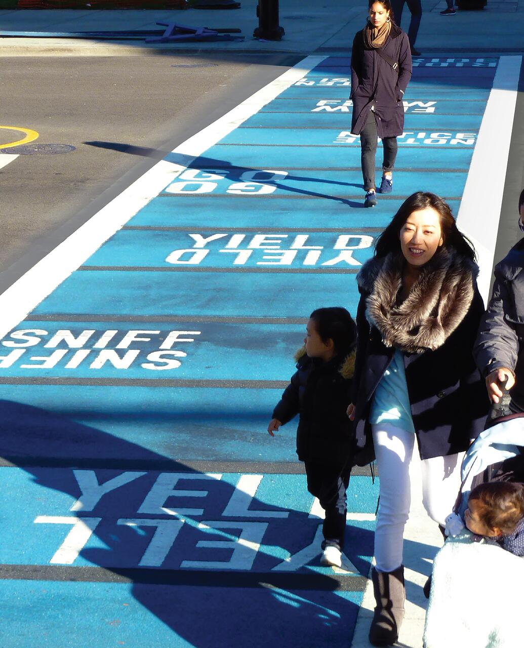 Pedestrians crossing a street painted with large blue and white text. A woman wearing a fur-trimmed coat is walking with a child in winter attire. Another person walks in the opposite direction. Shadows fall on the pavement.