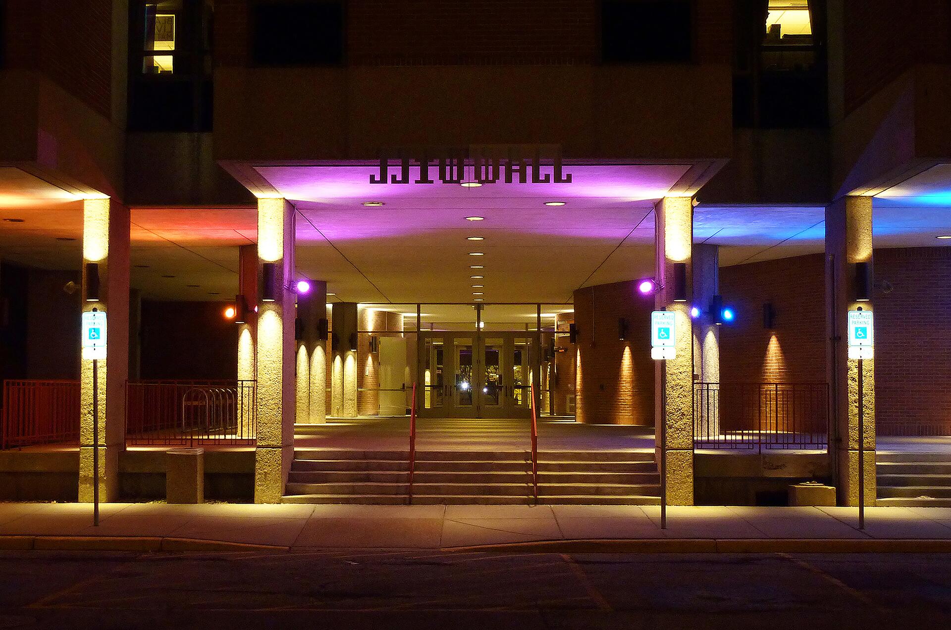 Building entrance at night with colorful lights illuminating columns, casting a warm glow on the steps and walls. Handicap parking signs are visible on either side.