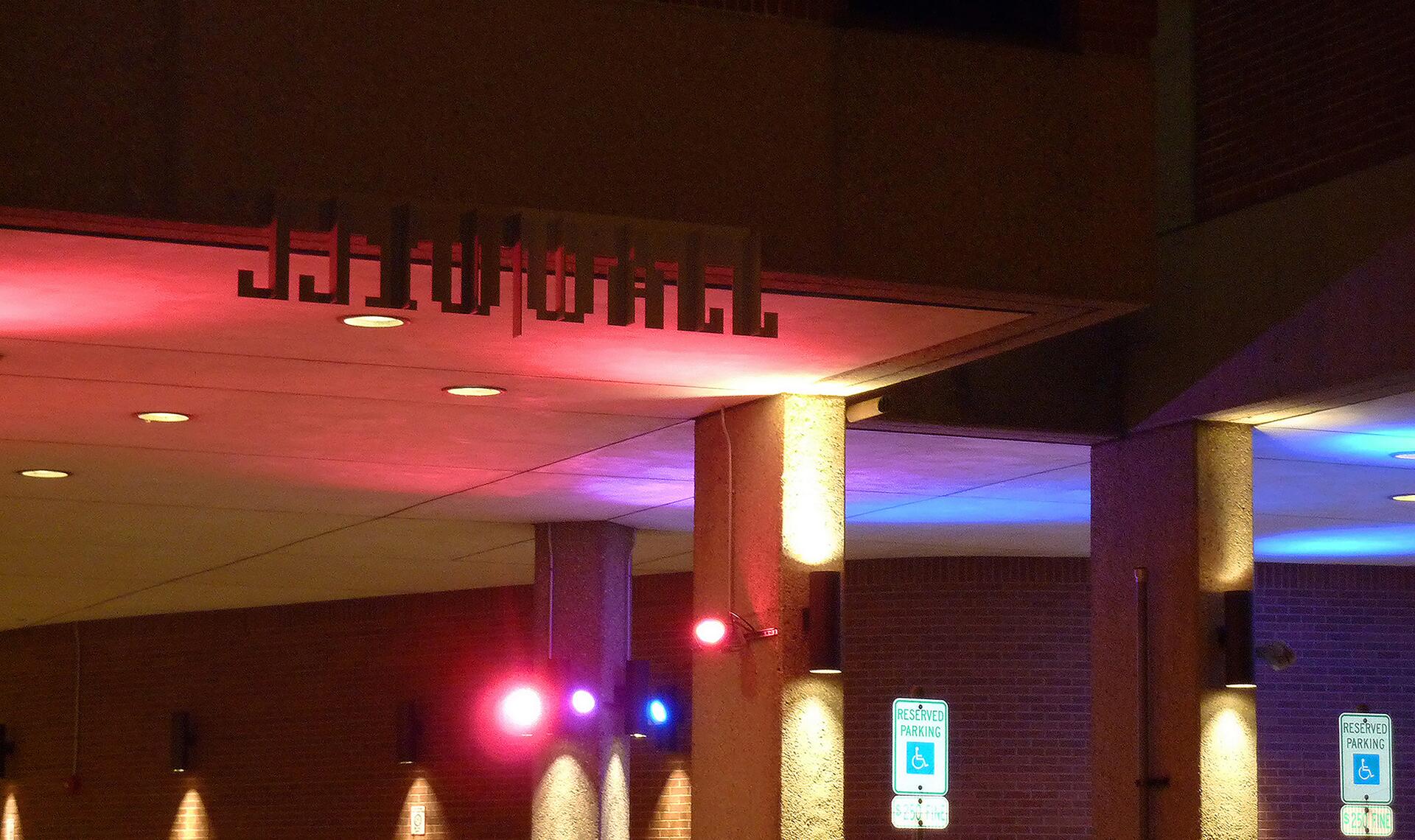 Colorful lights cast red, blue, and purple hues under an overhang with a silhouetted sign. Two handicap parking signs are visible below. The setting is night with warm and vibrant lighting.