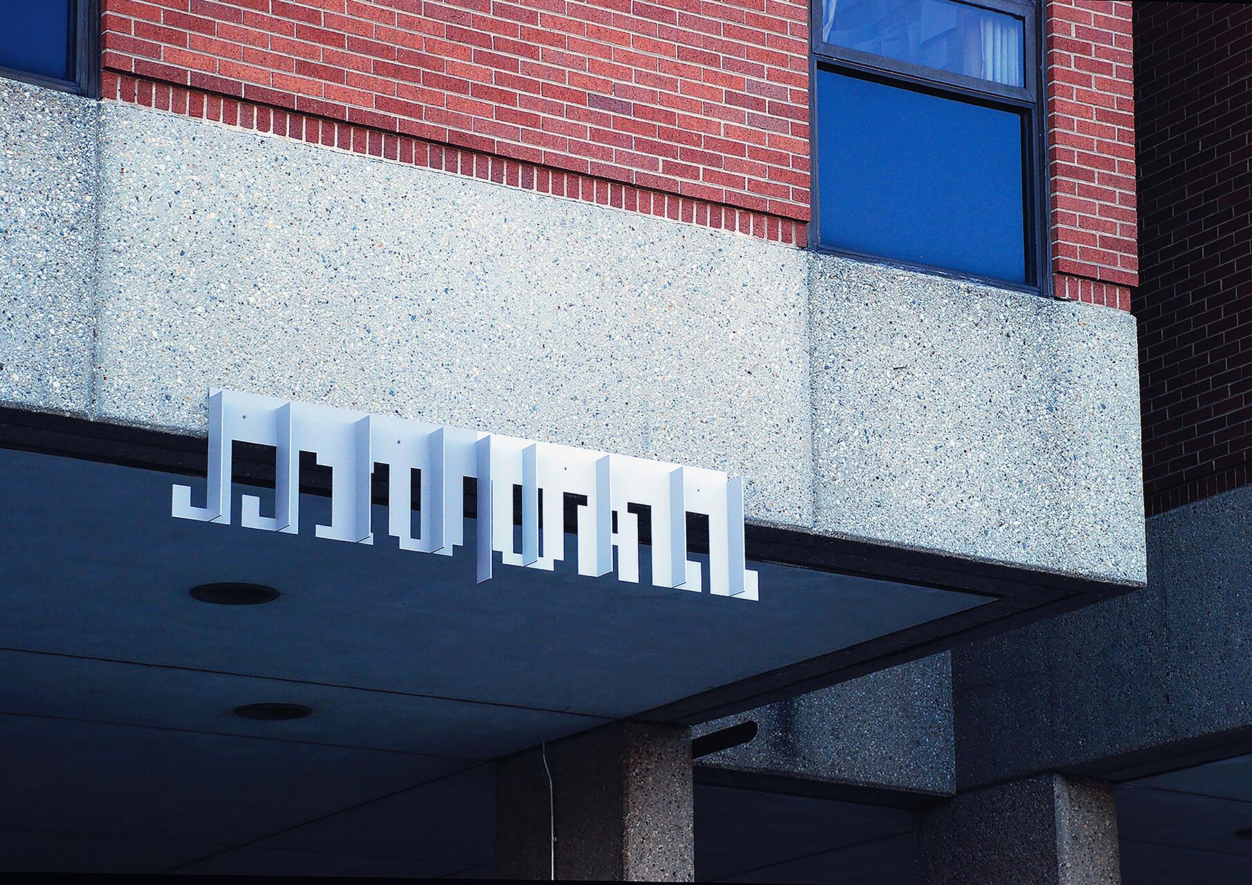 Building facade with a modern, abstract, white 3D sign extending from a concrete overhang against a backdrop of red brick and a blue-tinted window.