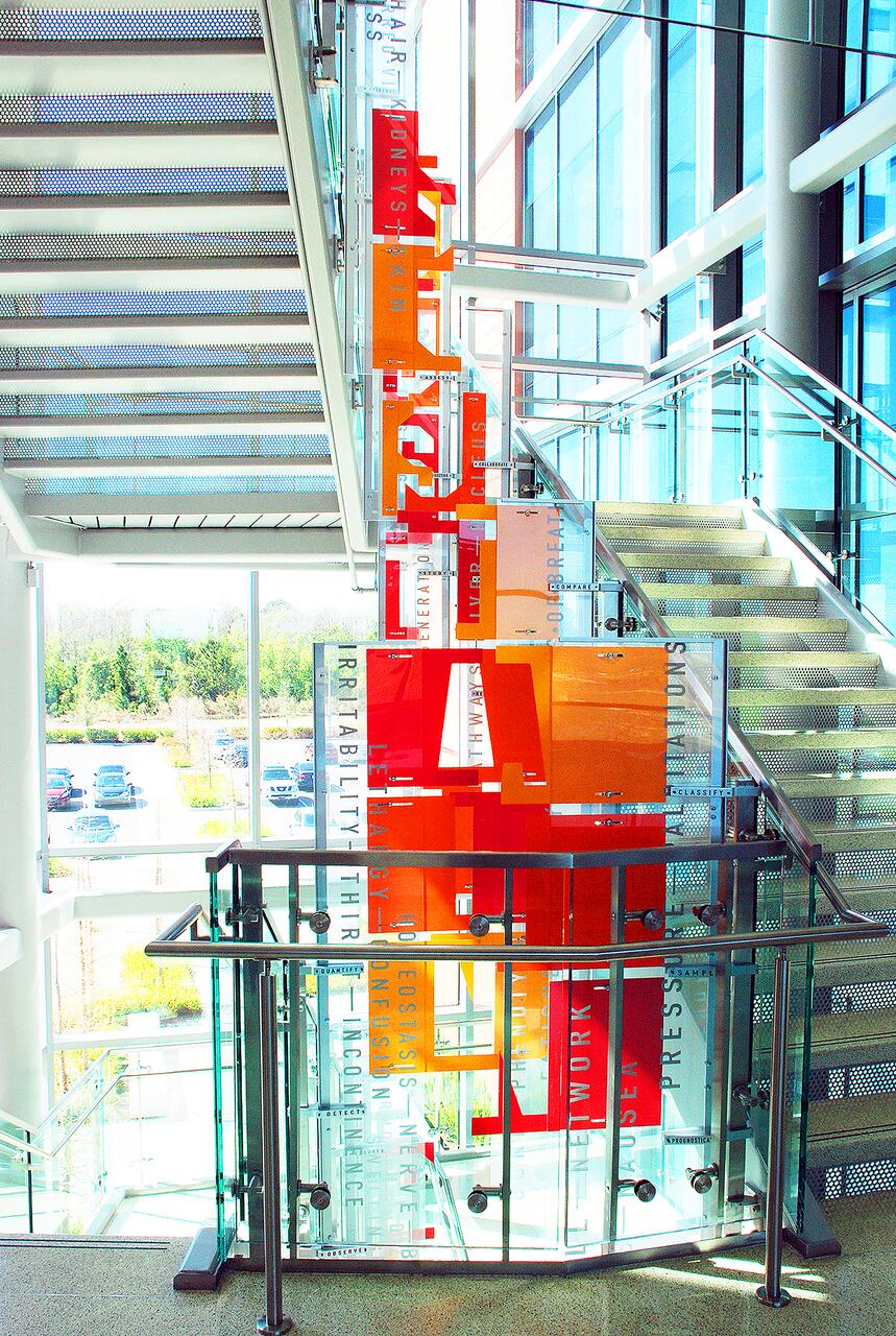 Colorful modern staircase with orange and red geometric panels, surrounded by glass and steel railings, in a bright building. Black text overlays some panels, with a view of parked cars and greenery through large windows.
