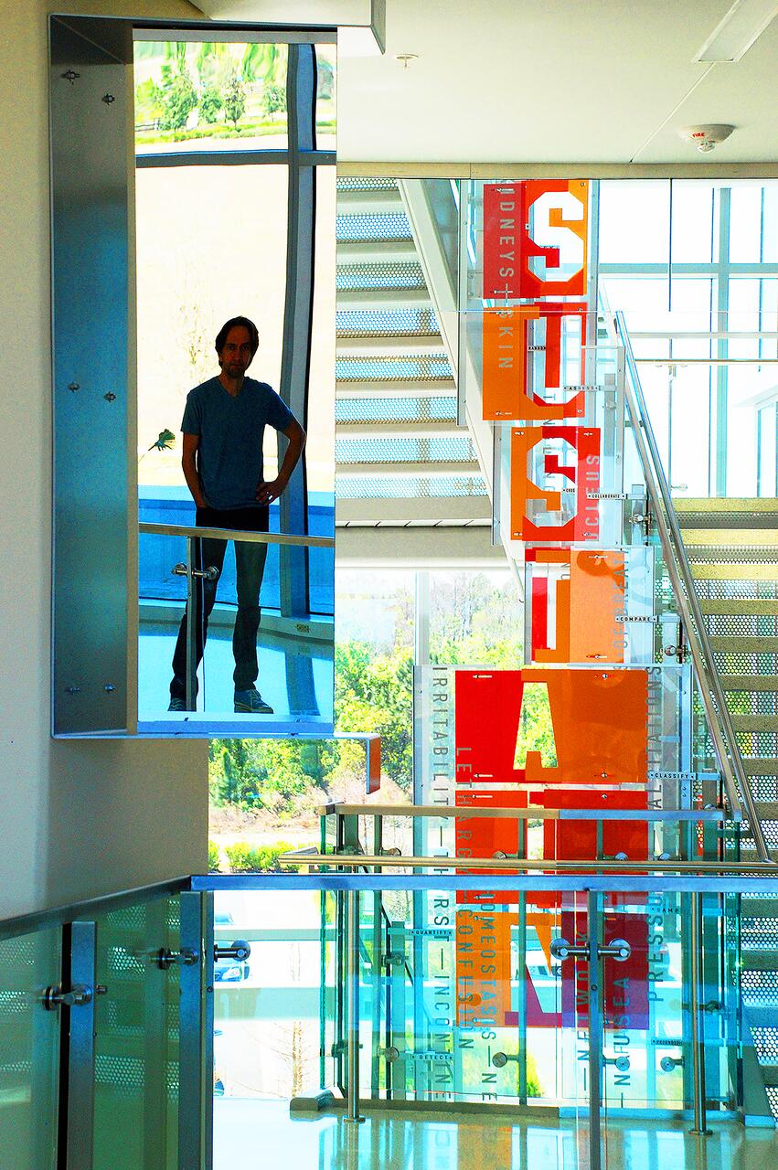 Man standing in front of a reflective panel in a modern, bright interior with glass railings and colorful signage. A staircase leads upward, and outside greenery is visible through large windows.