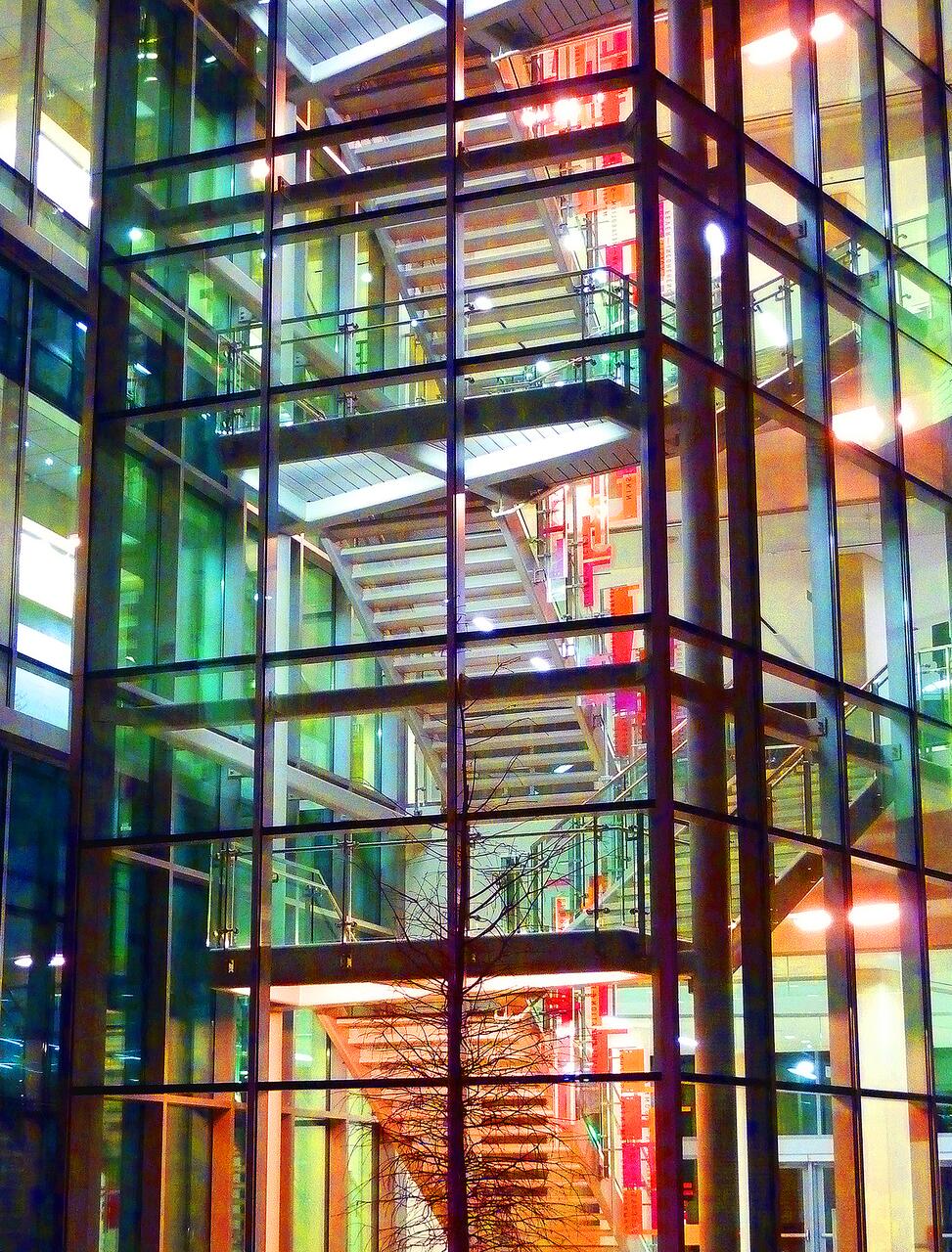 Glass-walled building with illuminated interior stairs. Bright colors from lights and reflections create a vibrant effect. A bare tree stands outside, contrasting with the modern architecture.