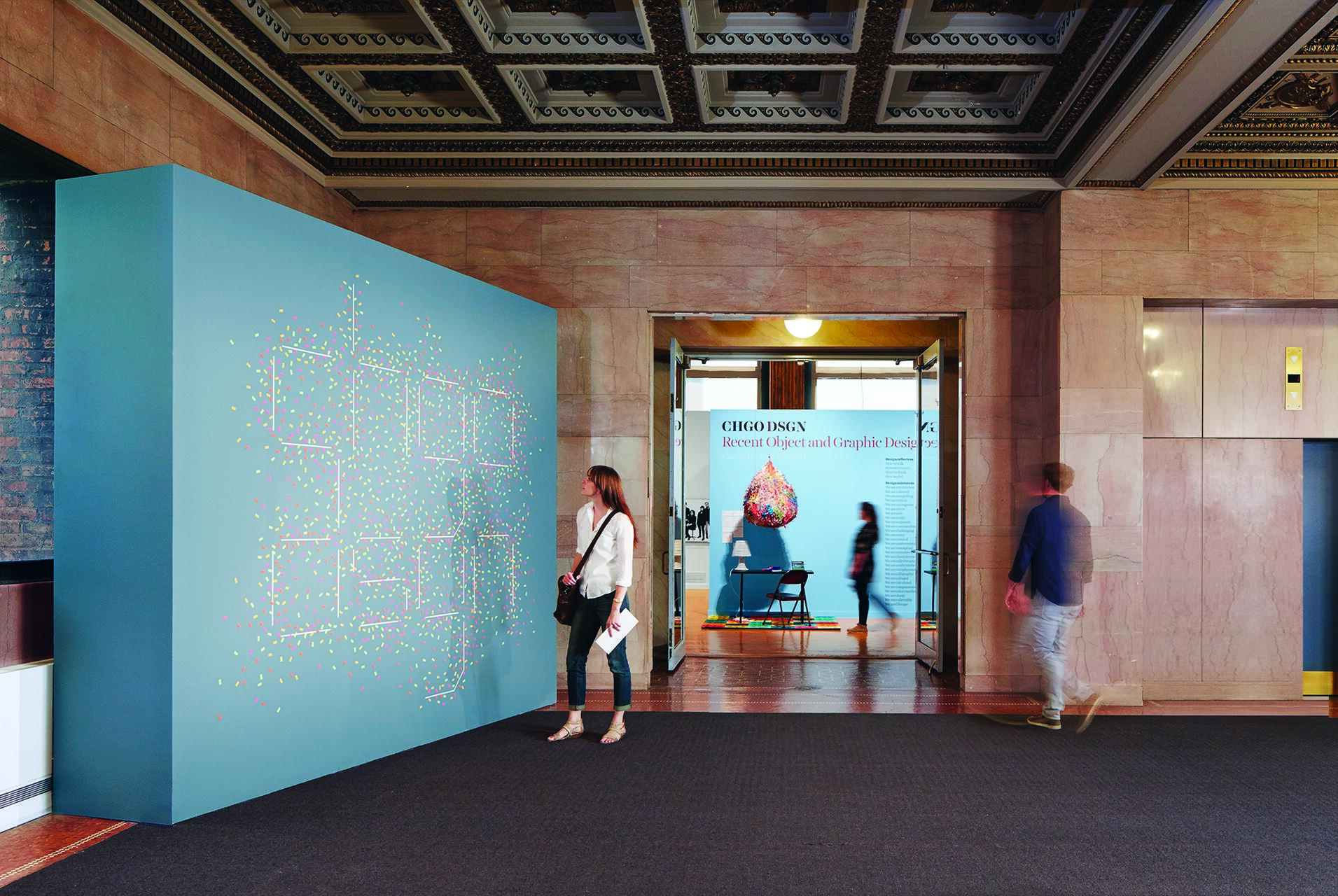 People walking in an exhibition hall, with a large blue wall on the left covered in colorful dots. A woman stands observing. The room in the background has a colorful display and text on a blue wall.