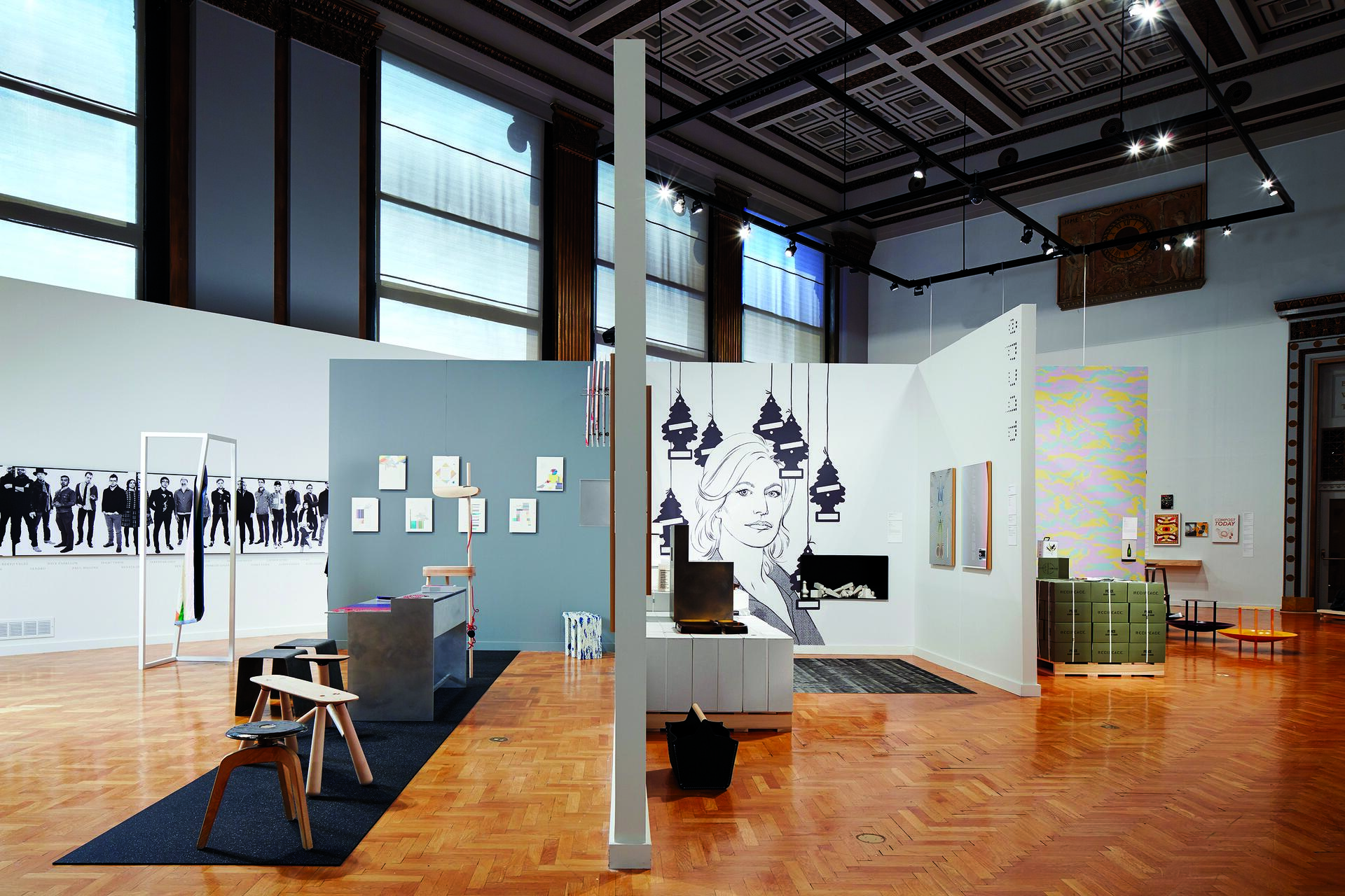 Gallery exhibition with modern art pieces, including framed art on walls, abstract installations, and a large drawing of a face. Wooden parquet flooring and high ceilings with large windows and track lighting.