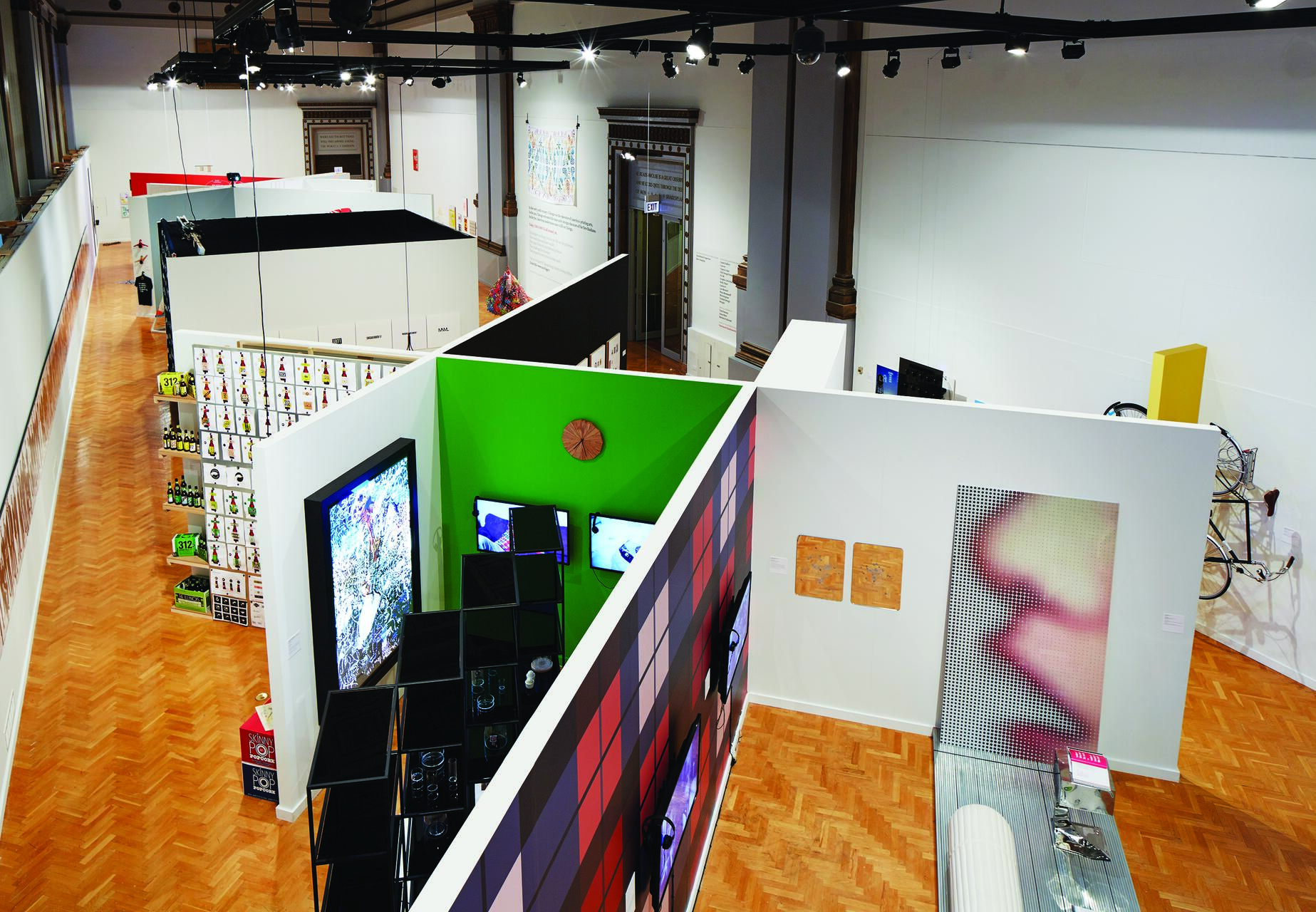 Art gallery with modern exhibits on patterned walls, wooden floors, and colorful displays. Various art pieces hang on white walls, including digital screens, a large pixelated face, and geometric patterns. Overhead lights illuminate the space.