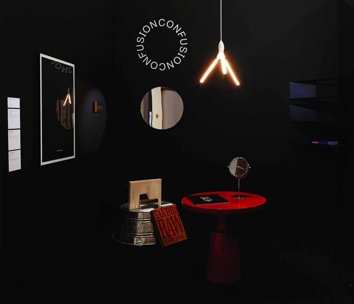Dark room with a red table, circular mirror, and hanging lights. Books and a red sign on a metal drum. A round mirror and shelves on the walls, with the word "CONFUSION" forming a circle above.