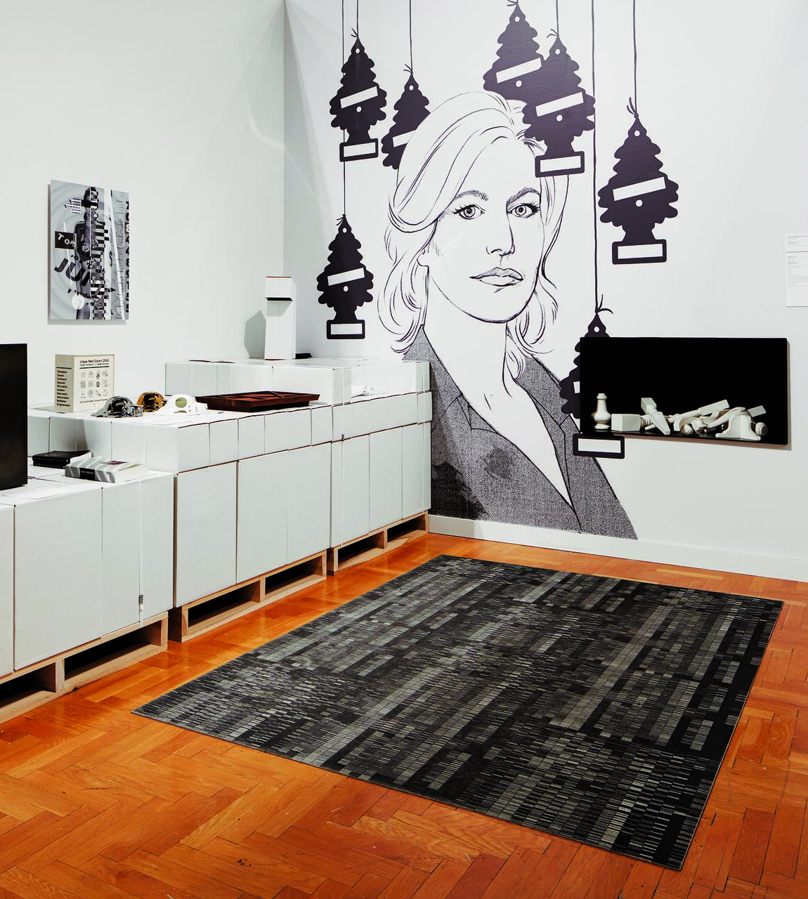 A room with white cabinets and a black-and-white mural of a woman on the wall, surrounded by hanging pine tree-shaped air fresheners. A black shelf holds small white objects. The floor has a patterned gray and black rug on wooden flooring.