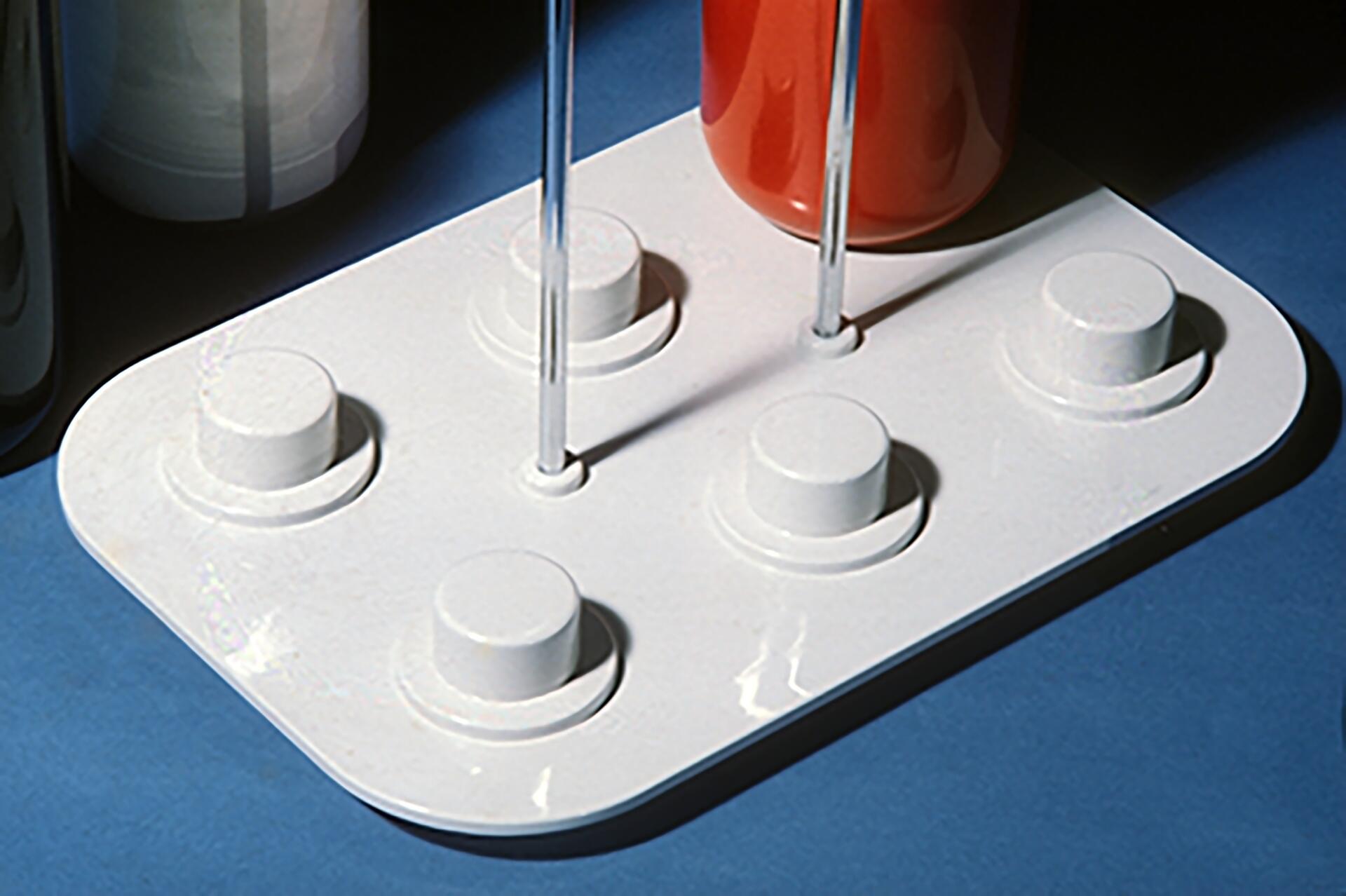 White base with six raised circular platforms, supporting a clear vertical rod, and an orange cylindrical object on a blue surface.