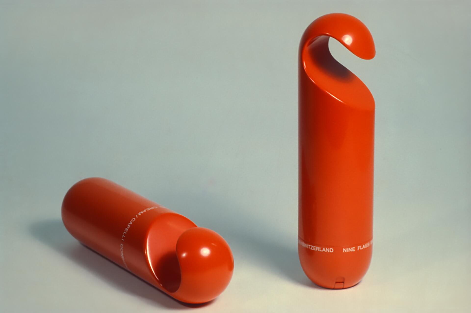 Two curved, elongated orange objects with a glossy finish against a light gray background. One is upright, showing a rounded hook-like top, while the other lies horizontally, revealing a spherical end. Subtle text visible near the base.