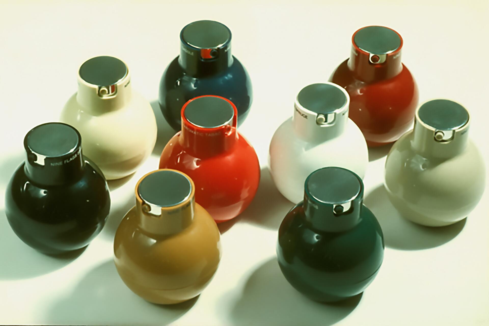 Nine colorful ceramic vases with round bases and flat tops arranged in a group. Colors include blue, black, red, white, beige, and brown. The vases have a modern, glossy finish and are placed on a light surface.