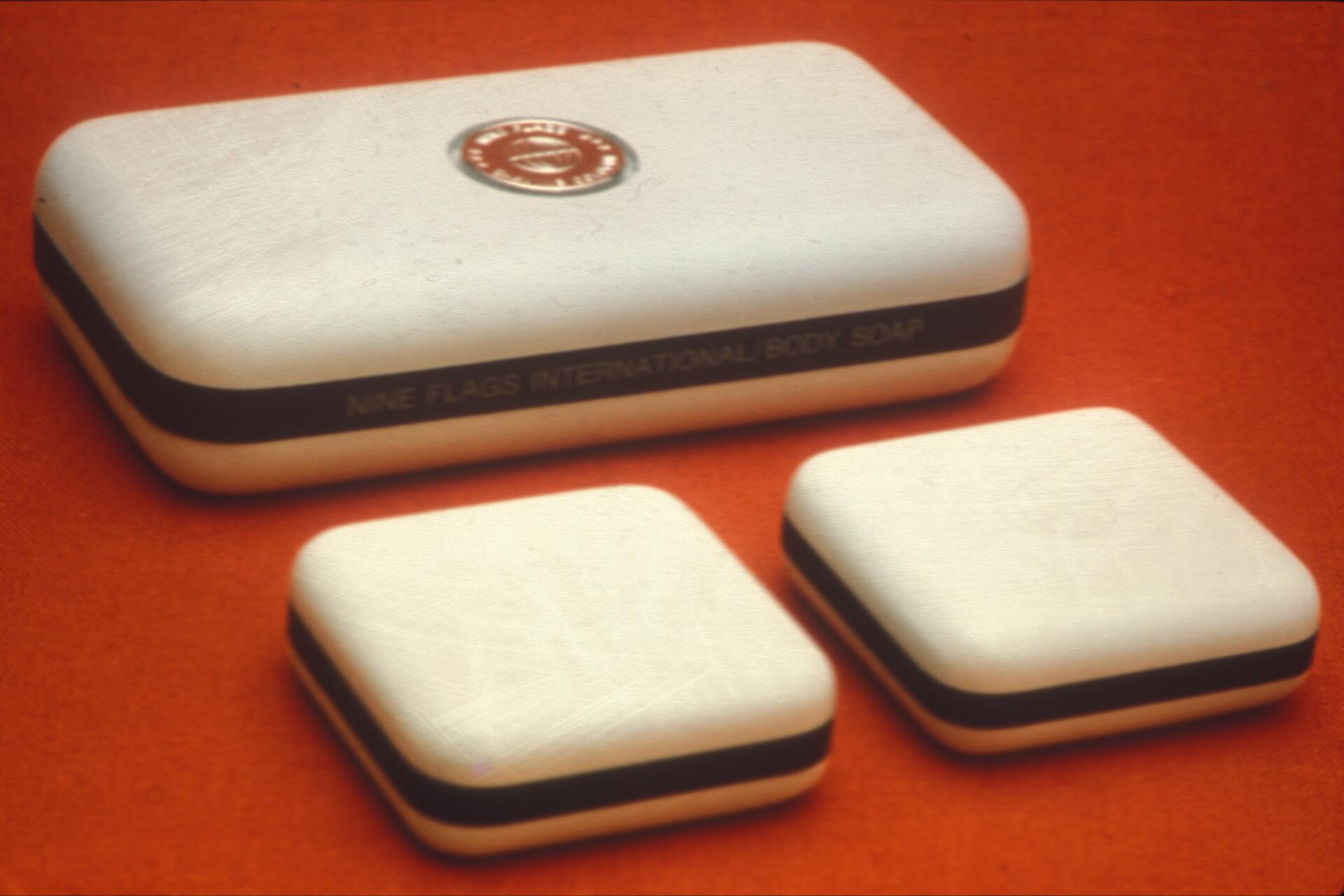 Three bars of white soap with black bands on an orange background. The largest bar shows text: "Nine Flags International Body Soap."